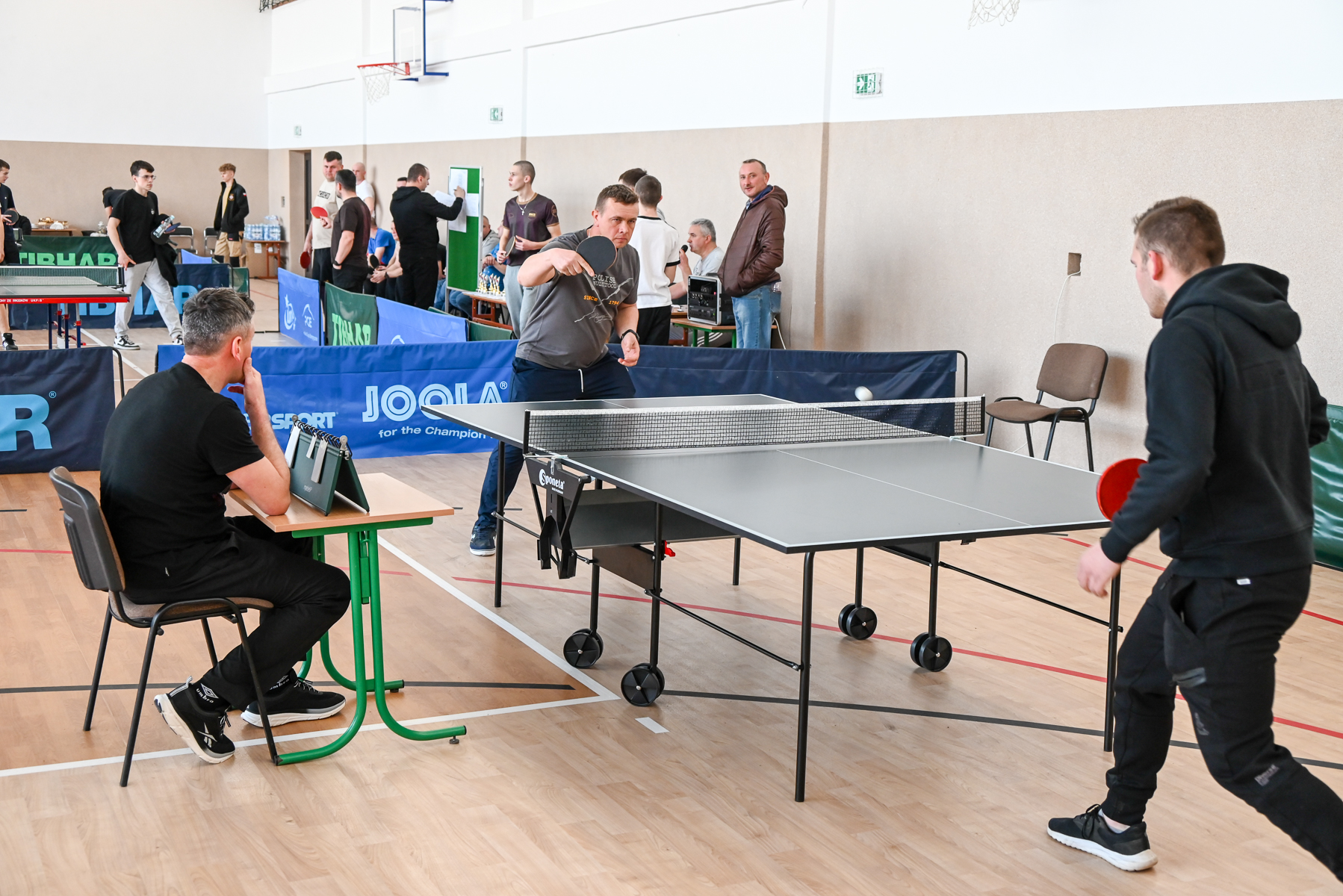 Mężczyźni grający w tenisa stołowego przy szarym stole w jasnej sali sportowej, sędzia siedzi przy stoliku z zielonym liczydłem, w tle osoby obserwujące i stojące przy ścianie.