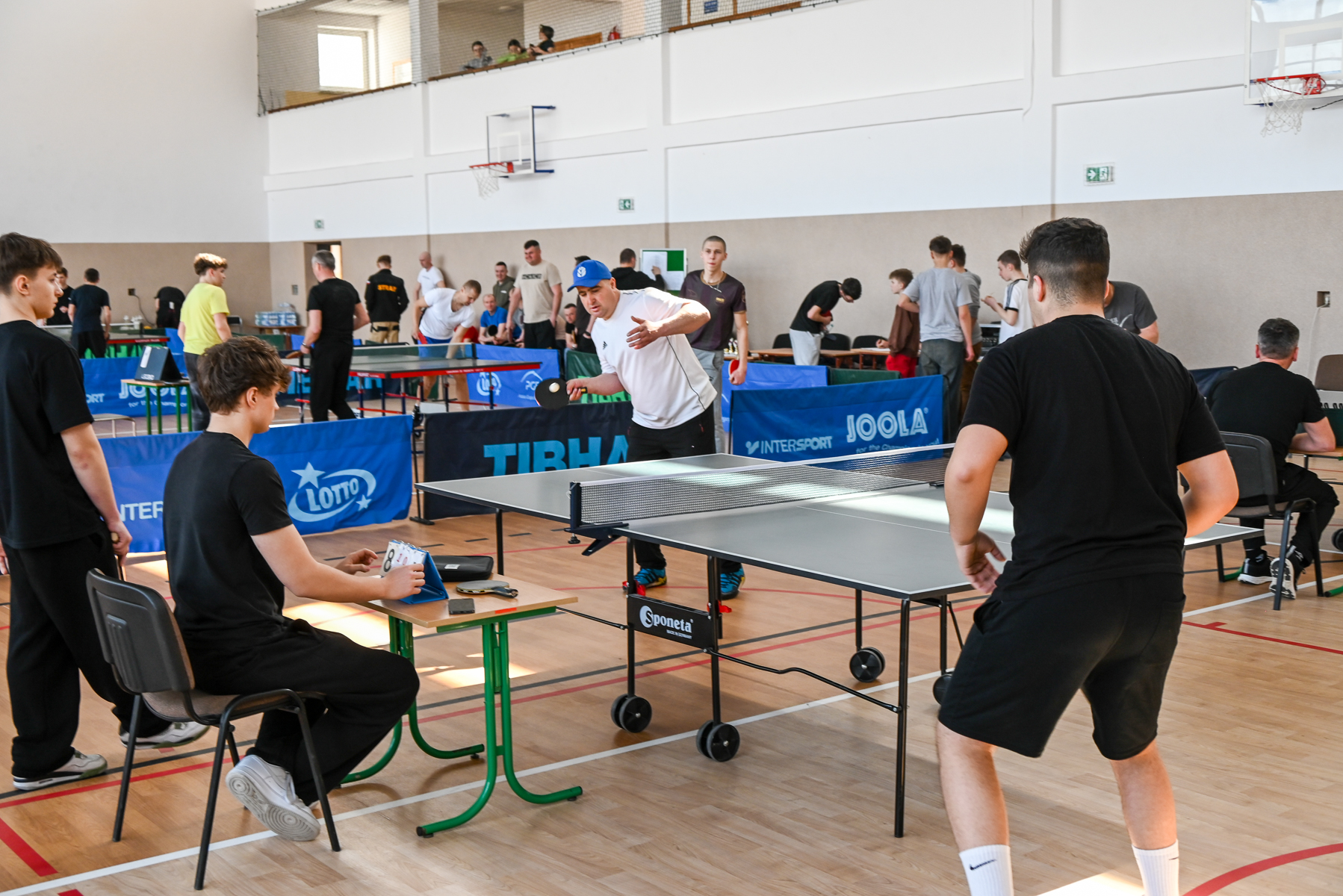 Hala sportowa z wieloma stołami do tenisa stołowego, zawodnicy i sędzia, ściany białe i beżowe, drewniana podłoga, niebieskie bandy reklamowe, kosze do koszykówki.