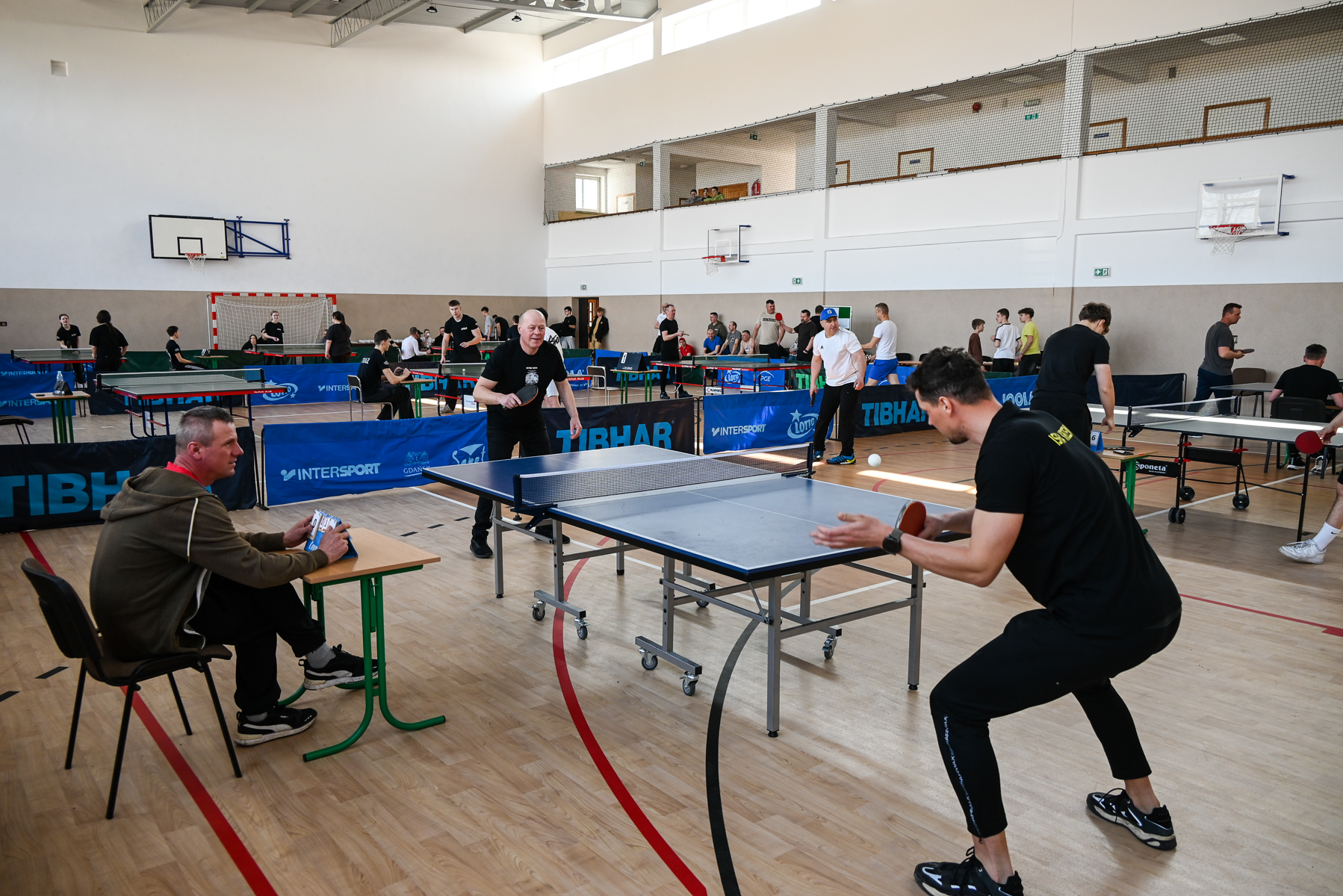 Hala sportowa z wieloma stołami do tenisa stołowego, gracze w ruchu, sędzia siedzący przy stole, drewniana podłoga, jasne ściany, naturalne światło.