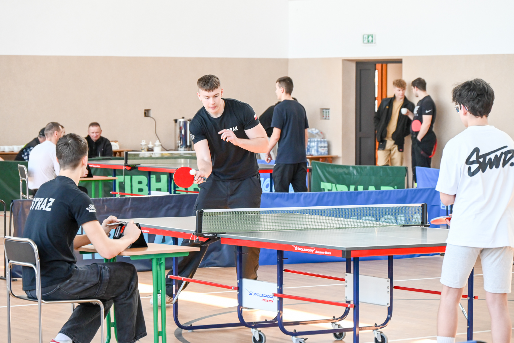 Młodzi mężczyźni grający w tenisa stołowego w hali sportowej, jeden z nich serwuje piłkę, sędzia z tabletem, w tle ludzie rozmawiający i siedzący przy stołach.