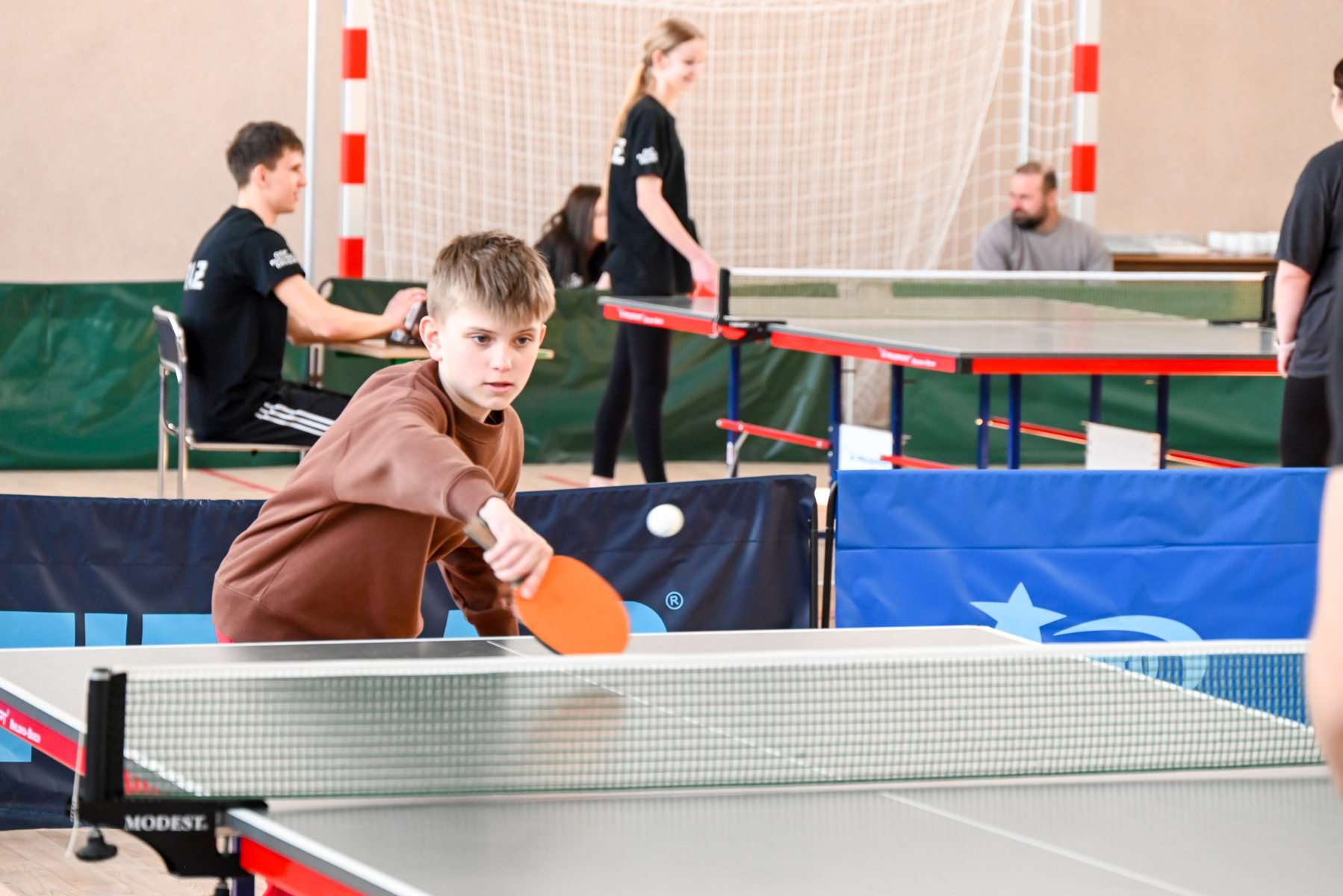 Chłopiec w brązowej bluzie uderza piłeczkę do tenisa stołowego na hali sportowej z innymi osobami w tle.