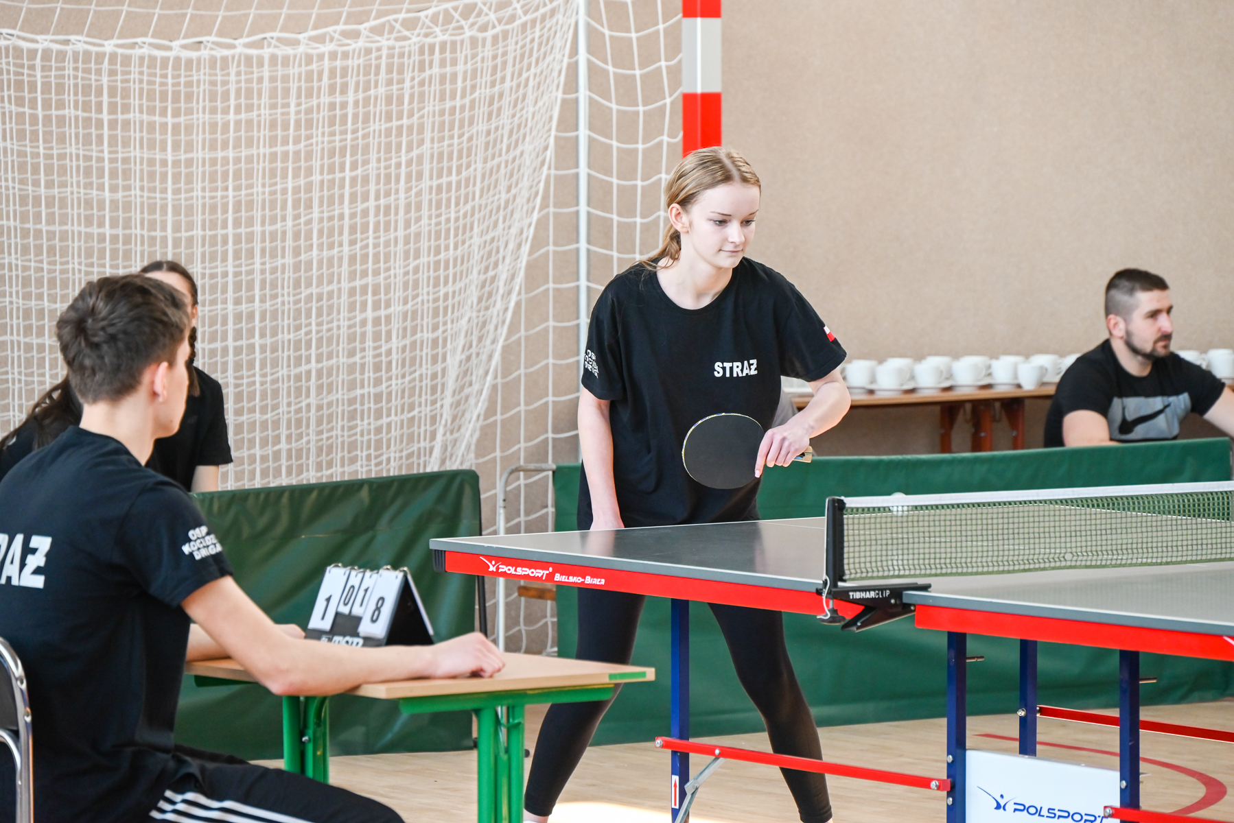 Dziewczyna w czarnej koszulce z napisem "STRAŻ" gra w tenisa stołowego przy stole z czerwoną ramą, mężczyzna siedzi przy stoliku z licznikiem punktów.