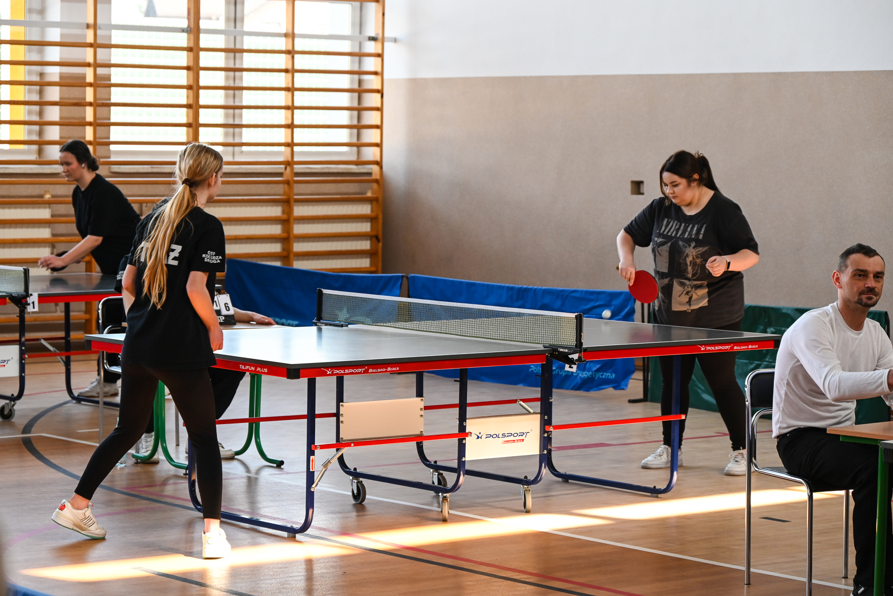 Cztery osoby w hali sportowej grające w tenisa stołowego, dwie kobiety przy stole w czarnych ubraniach, mężczyzna siedzący przy stole w białej koszulce, drewniana ściana gimnastyczna w tle.