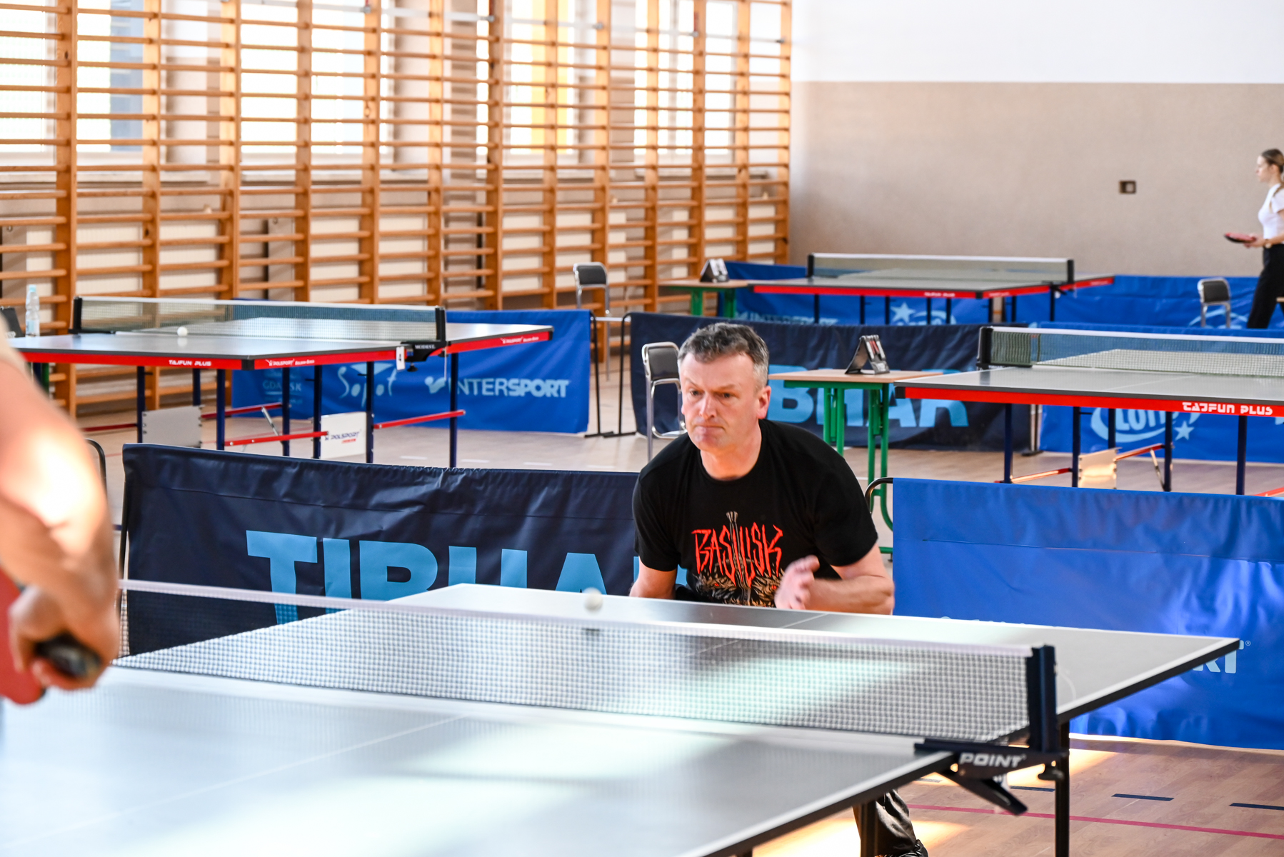 Mężczyzna w czarnej koszulce grający w tenisa stołowego w hali z drewnianymi drabinkami na ścianie, kilka stołów do ping-ponga i niebieskie barierki.