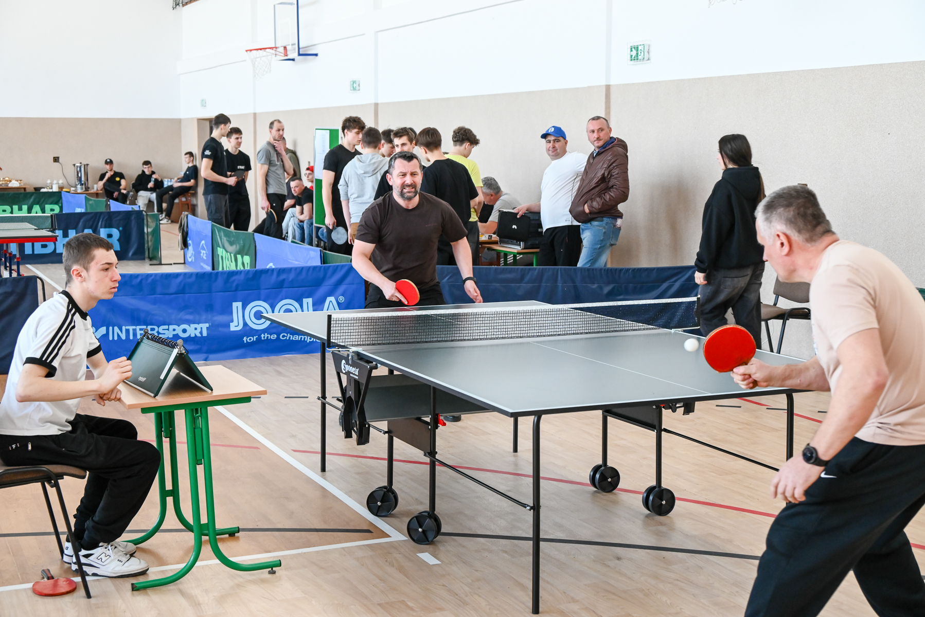 Dwóch mężczyzn gra w tenisa stołowego przy szarej składanej planszy, obok sędzia siedzi przy zielonym stoliku, w tle grupa ludzi stoi i siedzi w hali sportowej.