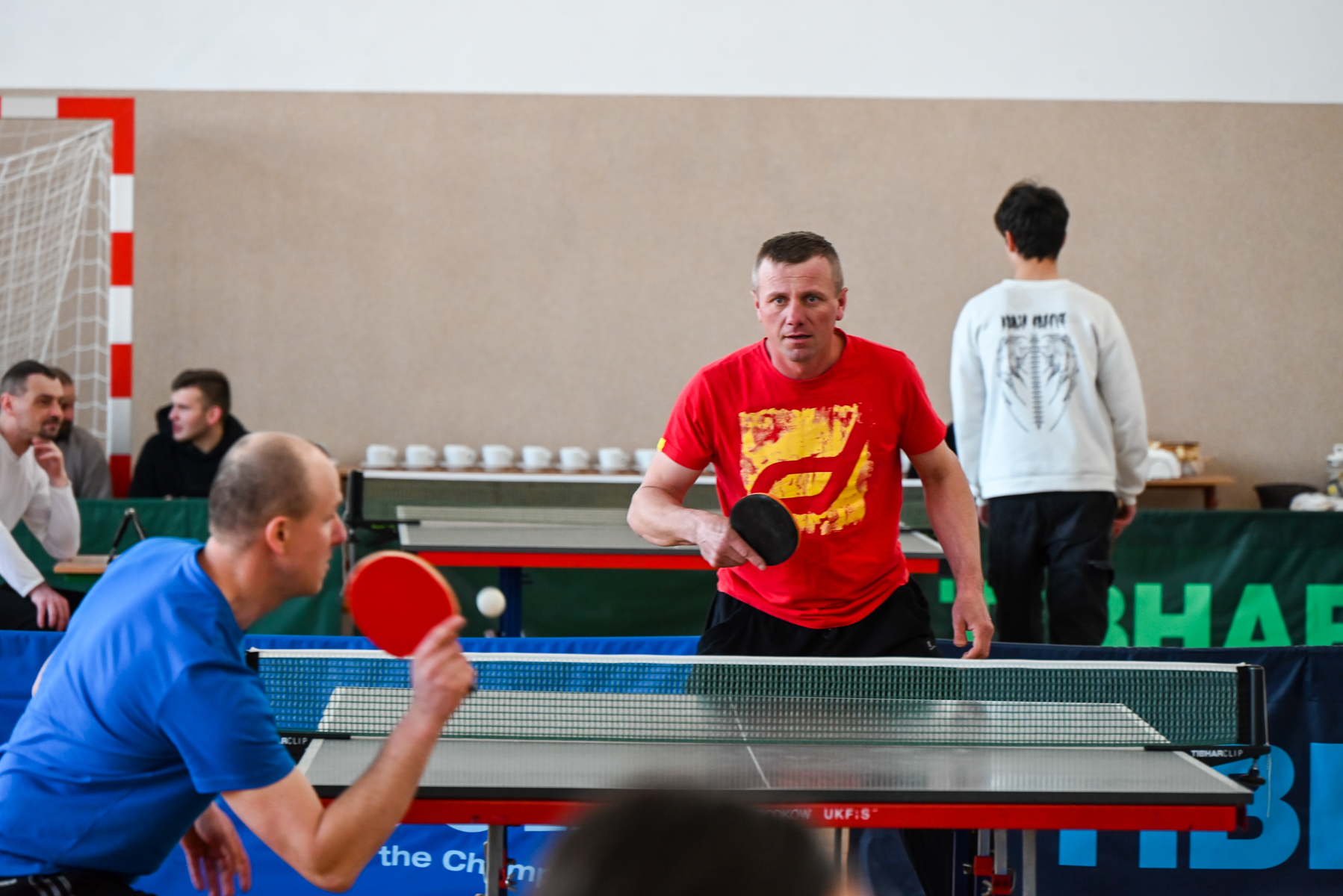 Dwóch mężczyzn grających w tenisa stołowego, jeden w niebieskiej koszulce, drugi w czerwonej, na tle stołu do ping-ponga i widowni.