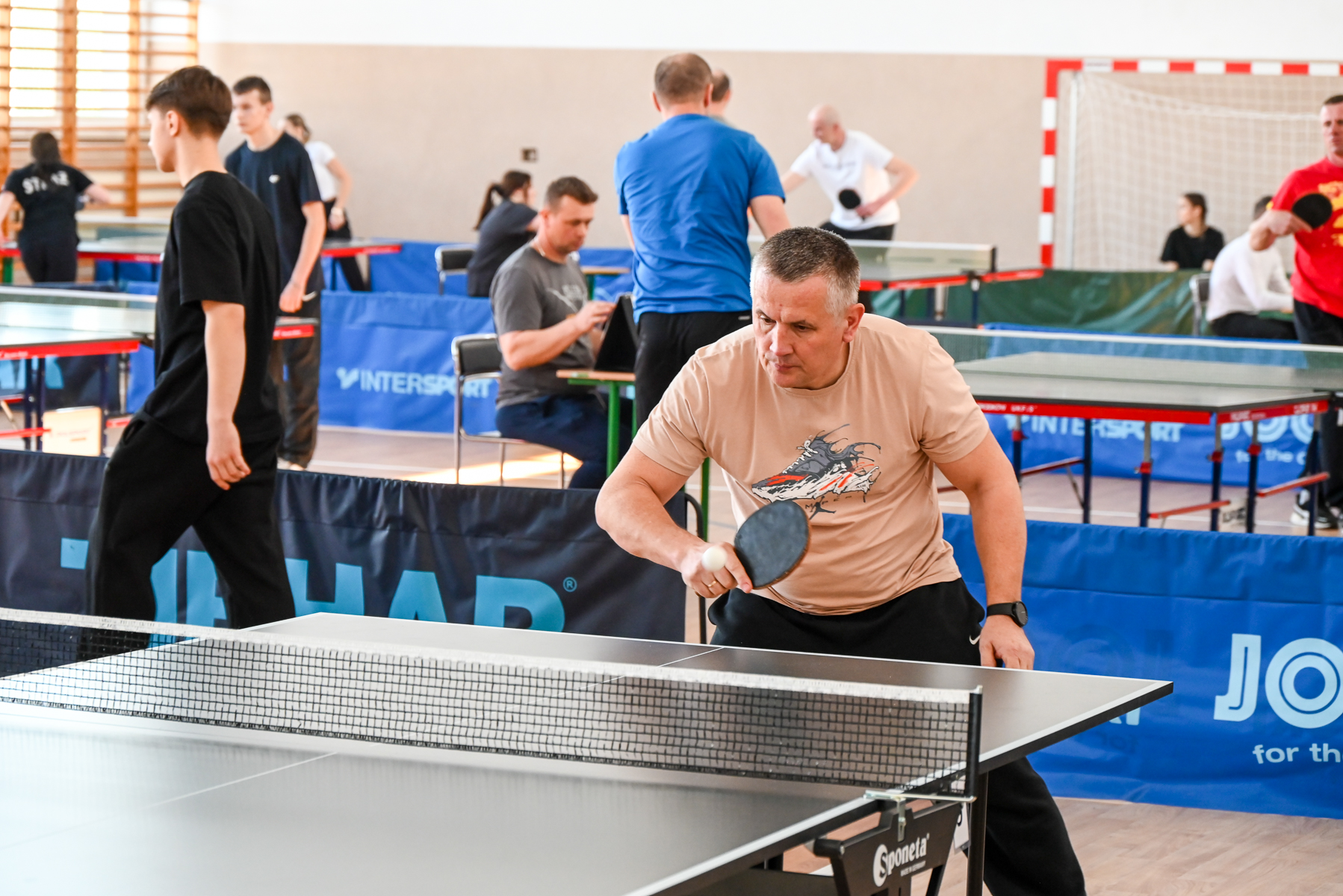 Mężczyzna w beżowej koszulce gra w tenisa stołowego na sali sportowej, w tle inni ludzie przy stołach do ping-ponga, niebieskie i czarne bariery, drewniana podłoga.