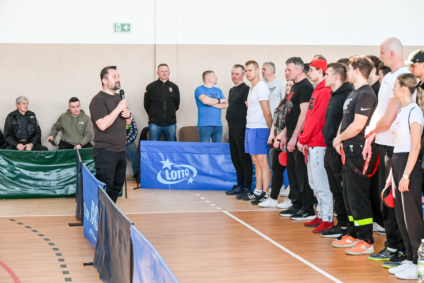 Mężczyzna z mikrofonem przemawia do grupy osób stojących w rzędzie na sali sportowej, kilka osób trzyma czerwone rakietki do tenisa stołowego.