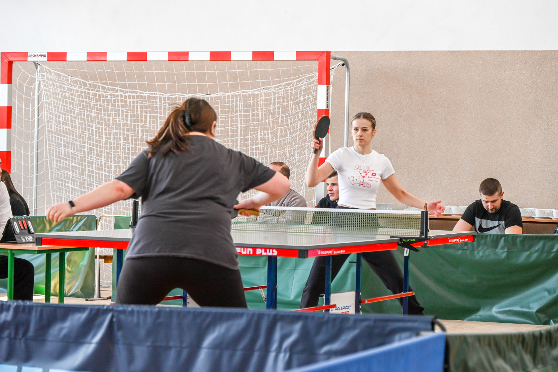 Dwie kobiety grające w tenisa stołowego przy stole z czerwoną ramą, za nimi bramka do piłki ręcznej z białą siatką, w tle osoby siedzące przy stole.