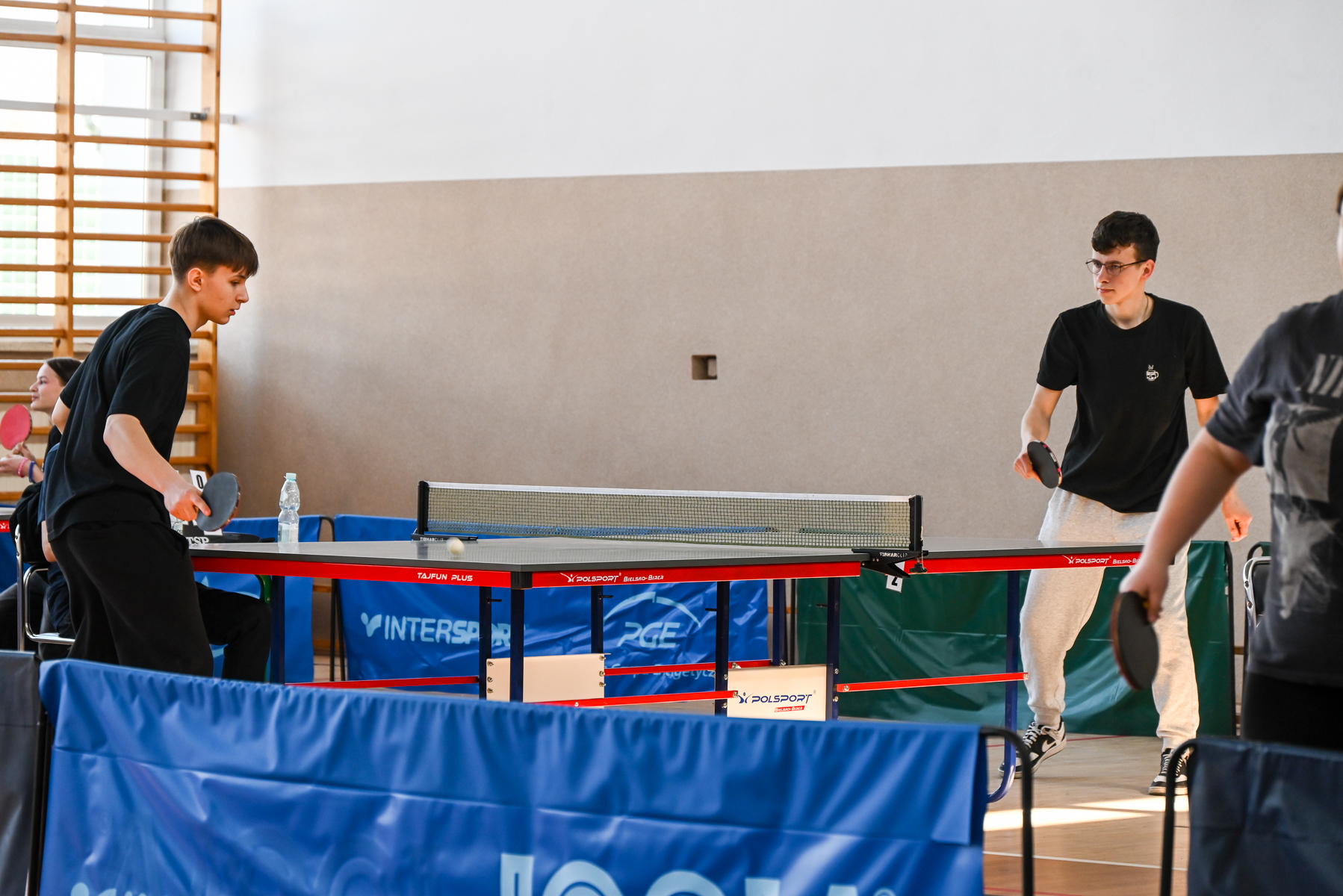 Dwóch chłopców grających w tenisa stołowego przy czerwono-czarnym stole, w tle jasna ściana i sportowa hala z drabinką.