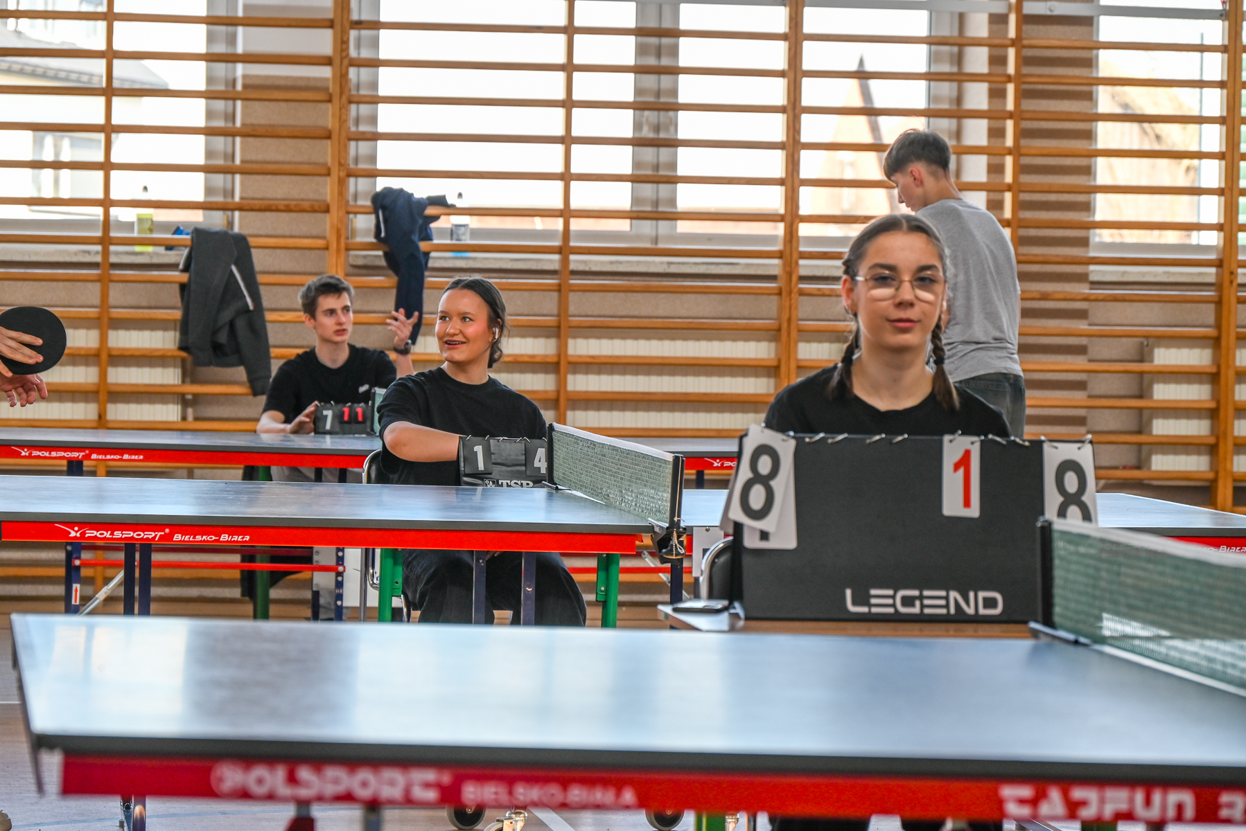 Trzy osoby siedzące przy stołach do tenisa stołowego, dwie z nich mają tablice wyników, drewniane drabinki na ścianie i duże okna w tle.