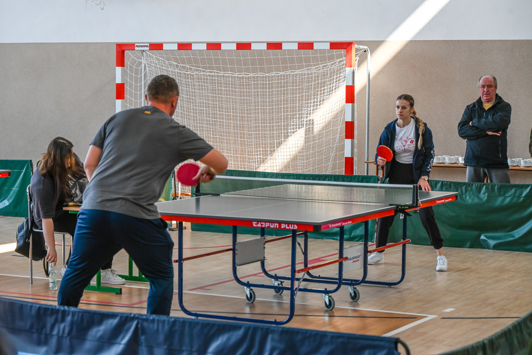 Dwóch ludzi gra w tenisa stołowego przy stole z czerwoną obwódką i zieloną siatką, w tle bramka do piłki ręcznej i cztery osoby obserwujące grę.
