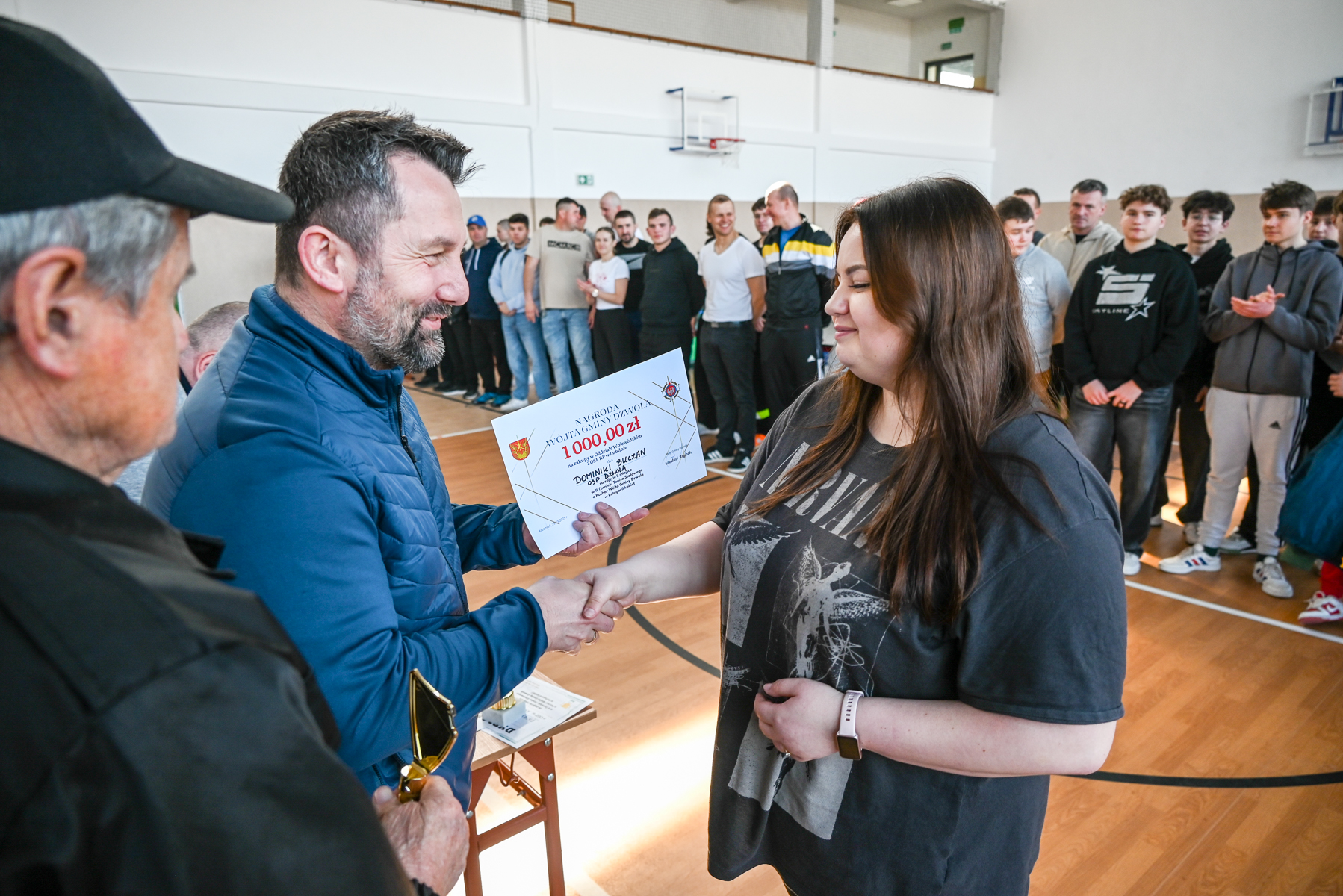 Mężczyzna w niebieskiej kurtce wręcza kobietę z długimi brązowymi włosami dyplom w hali sportowej, w tle stoi grupa osób na drewnianej podłodze.