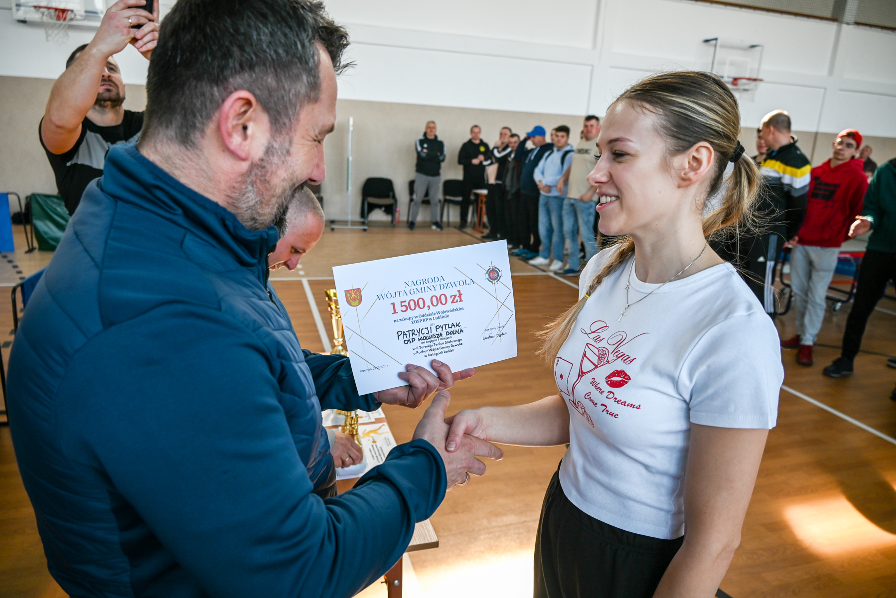 Mężczyzna w niebieskiej kurtce wręcza kobiecie z kucykiem i białą koszulką dyplom z napisem 1500 zł w hali sportowej, w tle stoi grupa osób.