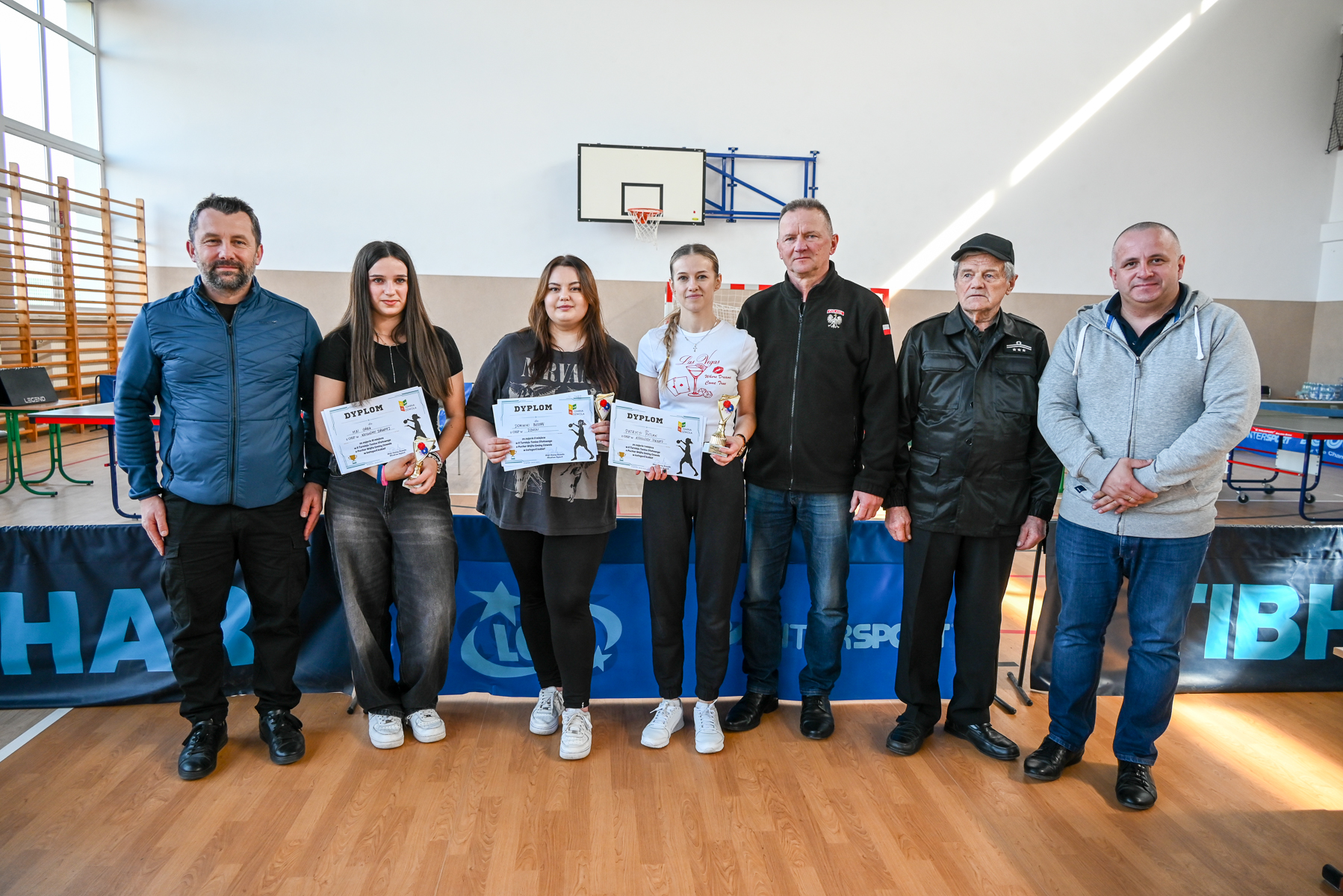 Siedem osób stoi w sali gimnastycznej na drewnianej podłodze, trzy kobiety w środku trzymają dyplomy i puchary, w tle kosz do koszykówki i siatka bramki.