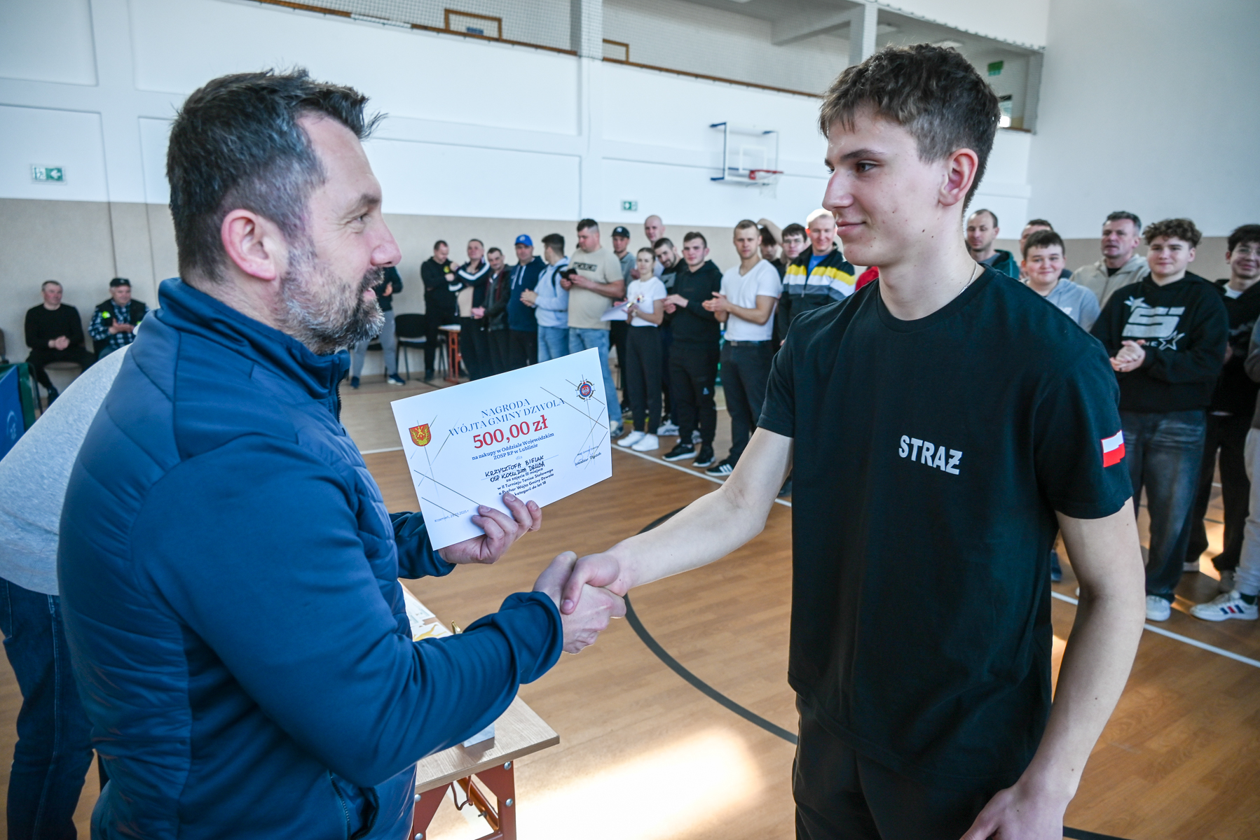 Dwóch mężczyzn ściska dłonie, jeden trzyma dyplom z napisem "500,00 zł", w tle grupa osób stojących na hali sportowej z drewnianą podłogą.