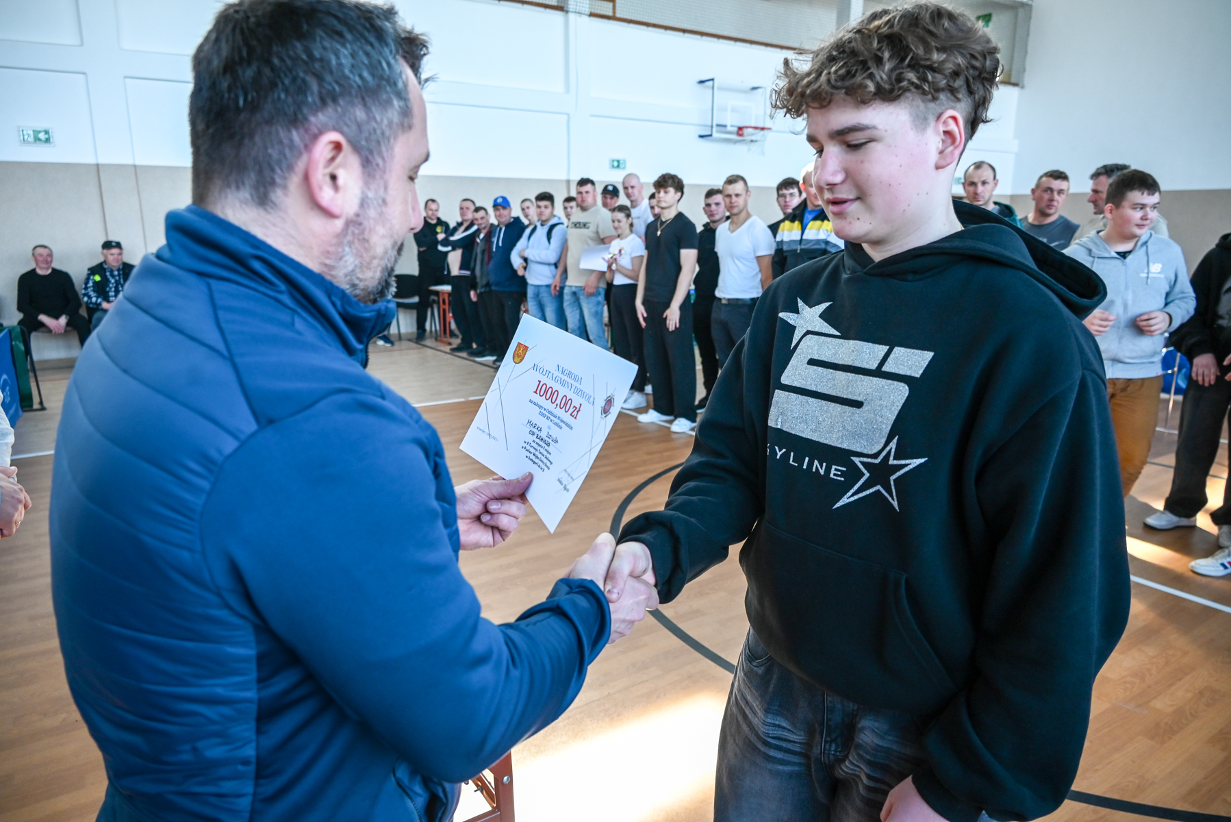 Dwóch mężczyzn ściska dłonie, jeden trzyma dyplom z napisem "1000,00 zł", w tle grupa osób stojących w hali sportowej z drewnianą podłogą.