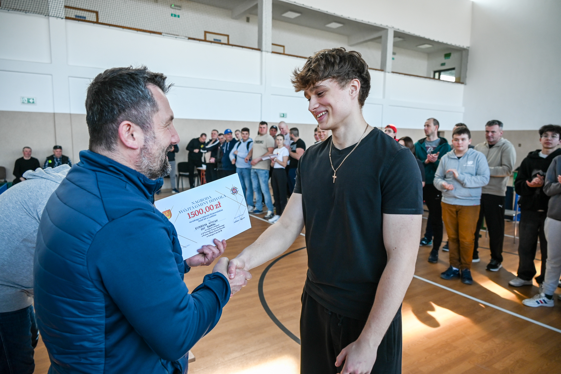 Dwóch mężczyzn ściska sobie dłonie, jeden trzyma dyplom z czerwonym napisem, w tle grupa ludzi klaszcze, wnętrze hali sportowej z jasnymi ścianami i drewnianą podłogą.