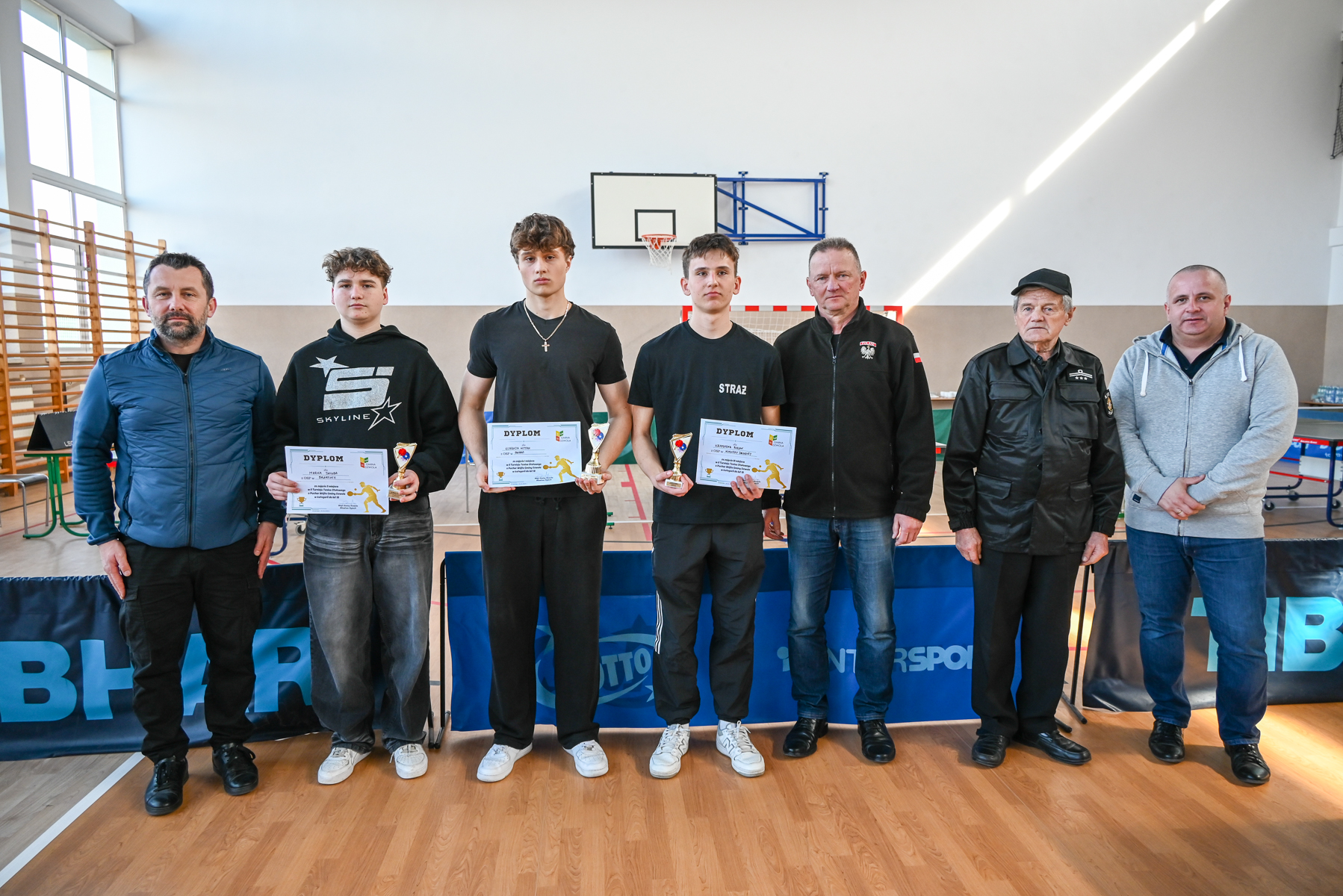 Siedem osób stojących na drewnianej podłodze hali sportowej, trzy środkowe osoby trzymają puchary i dyplomy, w tle bramka i tablica do koszykówki.