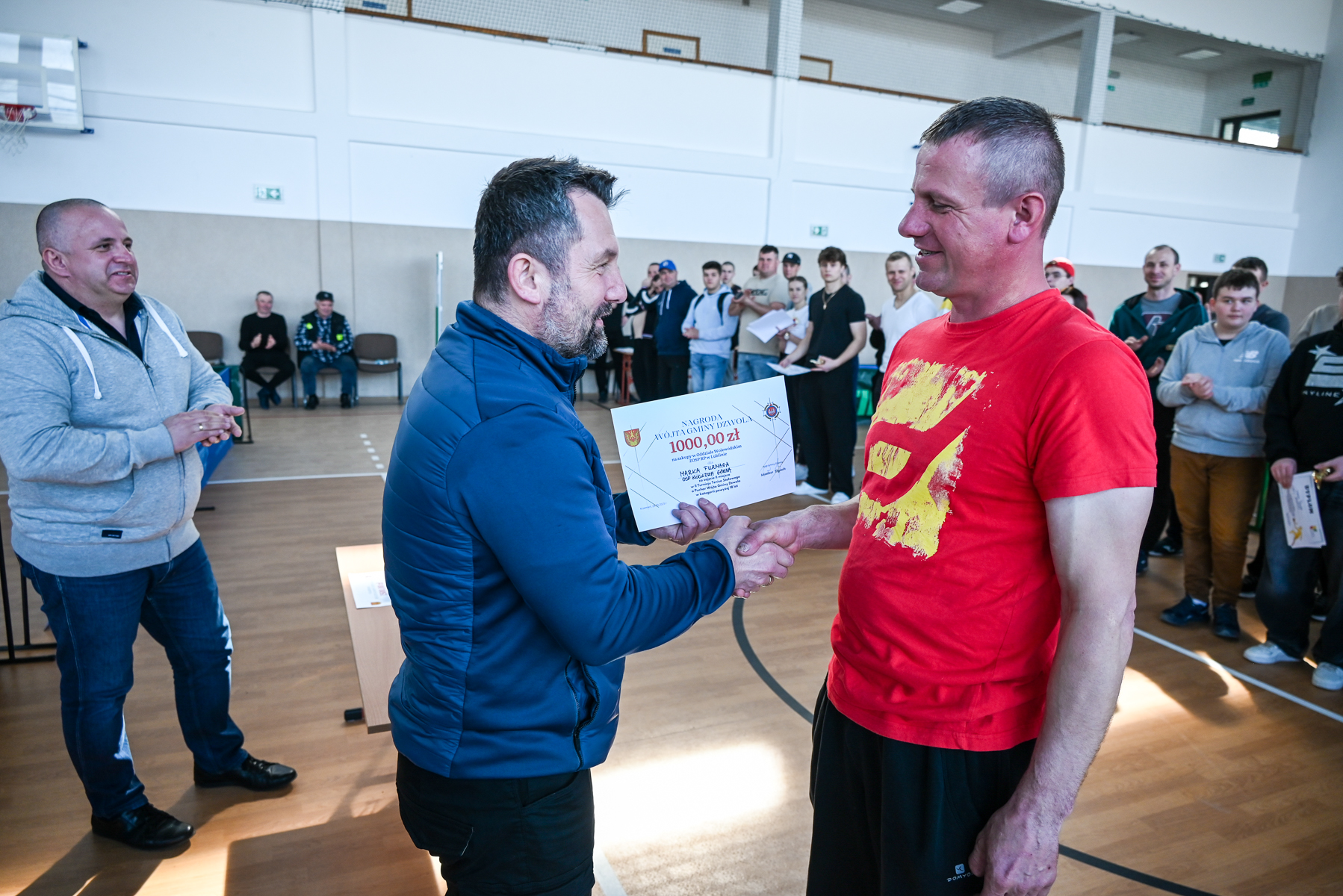 Dwóch mężczyzn podaje sobie ręce, jeden trzyma dyplom, w tle kilkanaście osób na sali sportowej z drewnianą podłogą i białymi ścianami.