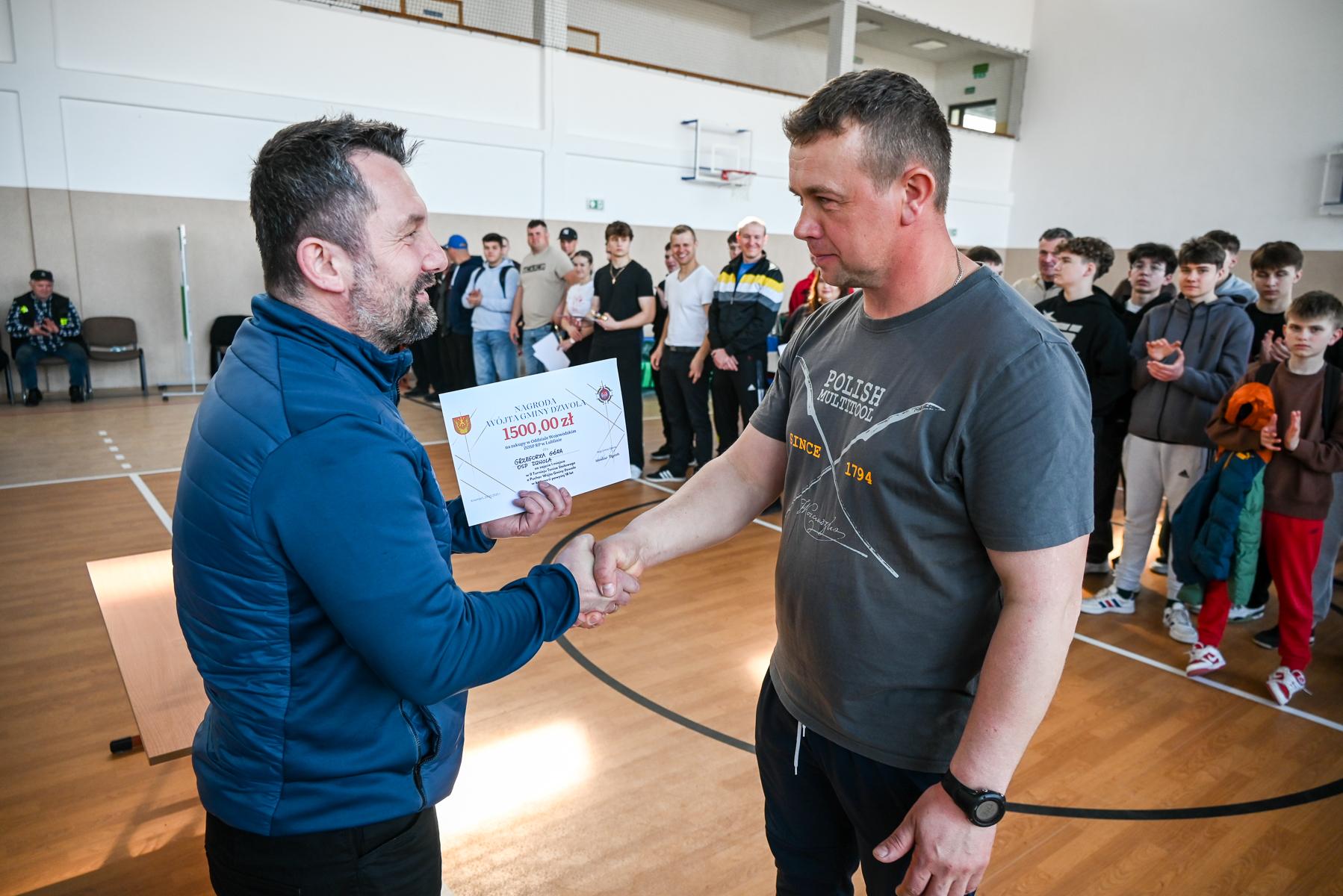 Dwóch mężczyzn ściska sobie dłonie, jeden trzyma dyplom, w tle grupa młodych ludzi, sala sportowa z drewnianą podłogą i ścianami w jasnych kolorach.