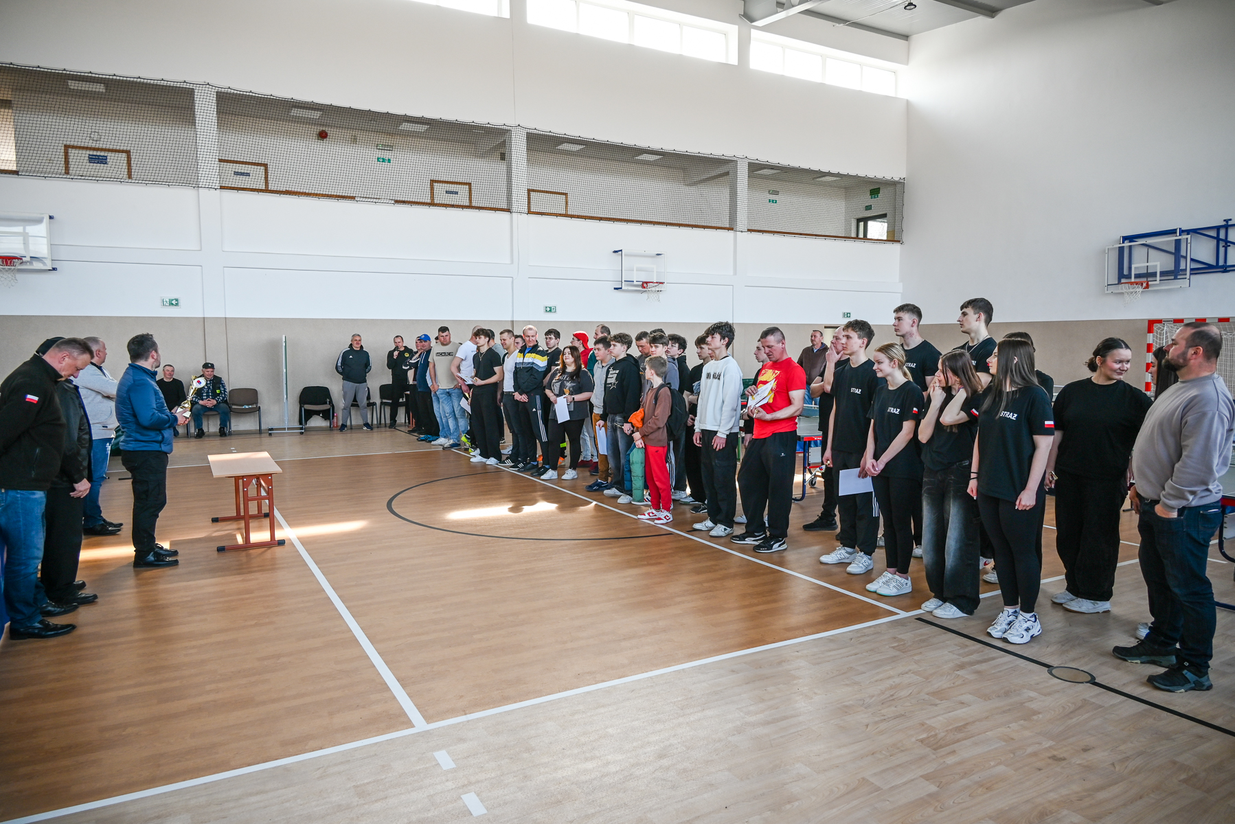 Grupa osób ustawiona w rzędzie na drewnianej podłodze hali sportowej, z lewej strony mężczyzna trzymający puchar, w tle ściana z oknami i koszami do koszykówki.