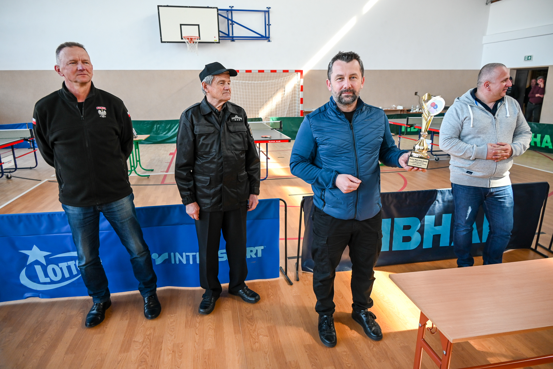 Czterech mężczyzn stoi na sali gimnastycznej, trzeci od lewej trzyma złoty puchar, w tle stół do tenisa stołowego i bramka.
