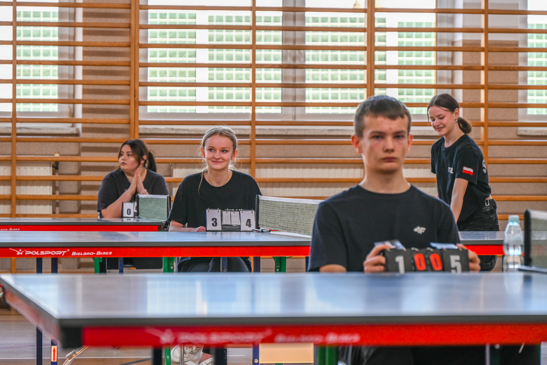 Cztery osoby w czarnych koszulkach przy stołach do tenisa stołowego w hali sportowej z drewnianymi drabinkami na ścianie, trzy liczniki punktów.