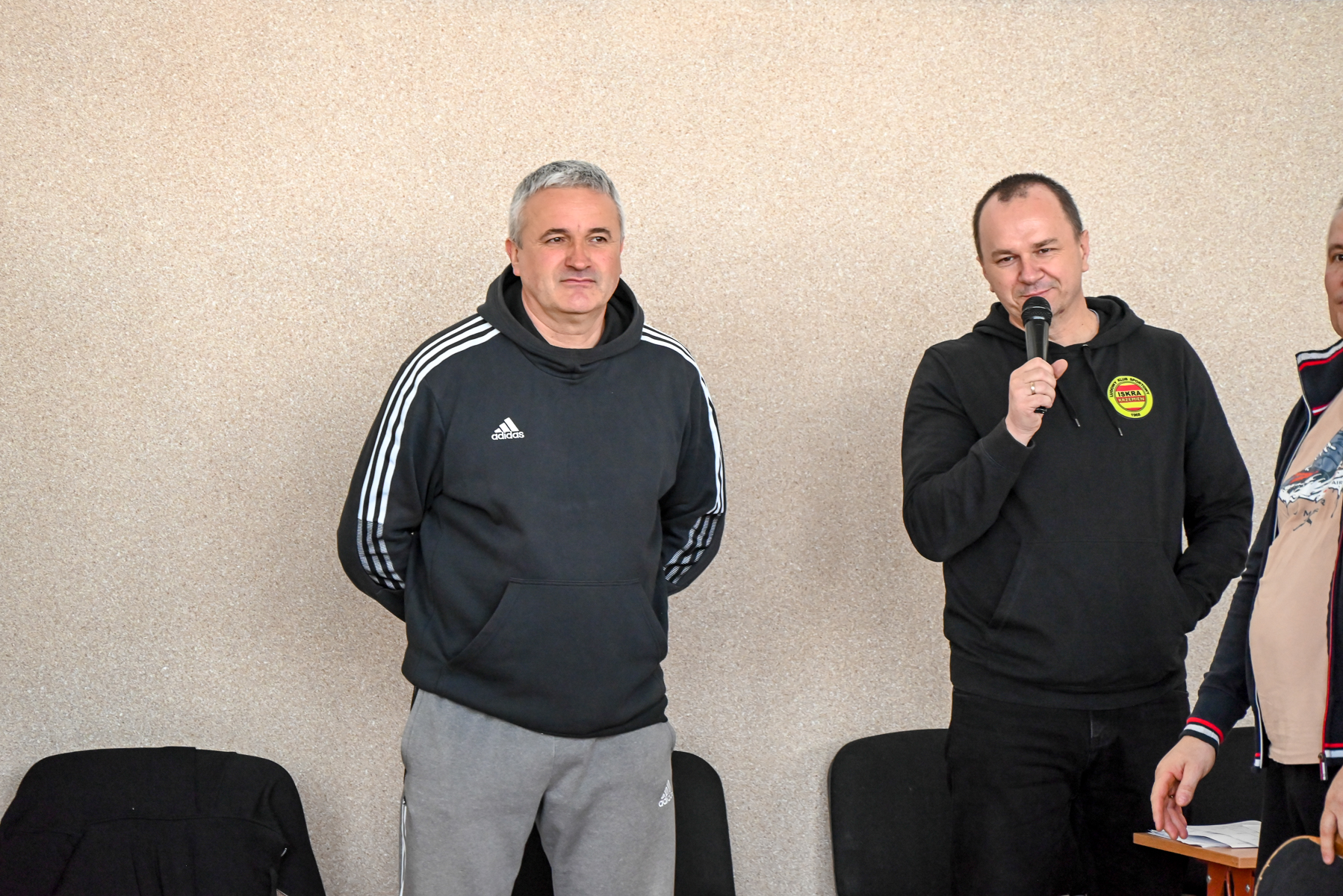 Dwaj mężczyźni w ciemnych bluzach z kapturem, jeden trzyma mikrofon, stoją na tle jasnej ściany, obok nich czarne krzesła i fragment stolika.