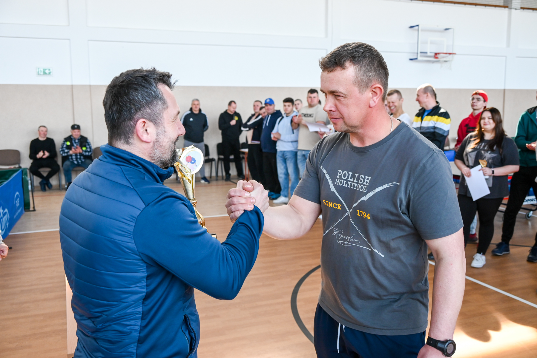 Dwóch mężczyzn ściska sobie dłonie, jeden trzyma złoty puchar, w tle ludzie stojący i siedzący na sali sportowej z drewnianą podłogą.