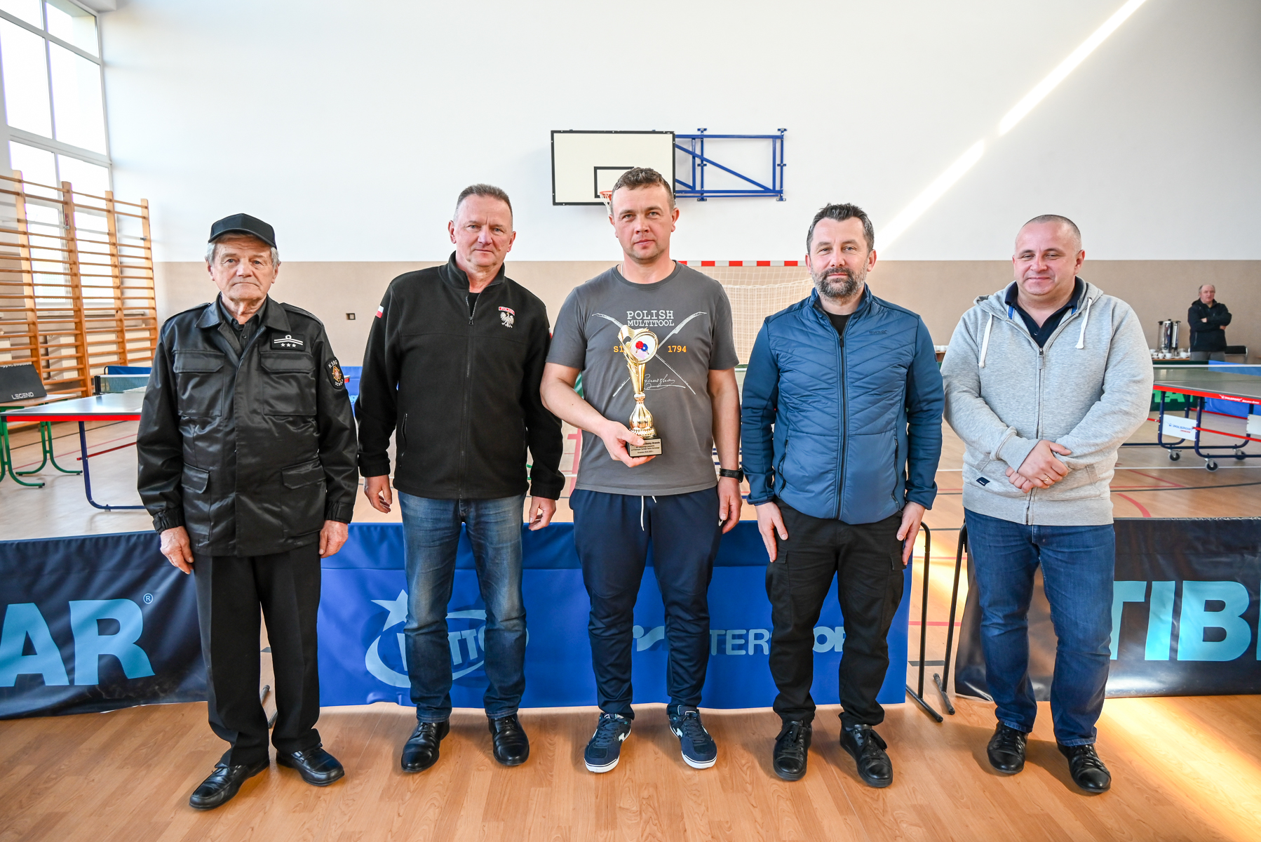 Pięciu mężczyzn stoi w hali sportowej, jeden w centrum trzyma złoty puchar, w tle bramka i kosz do koszykówki, jasne ściany, drewniana podłoga.