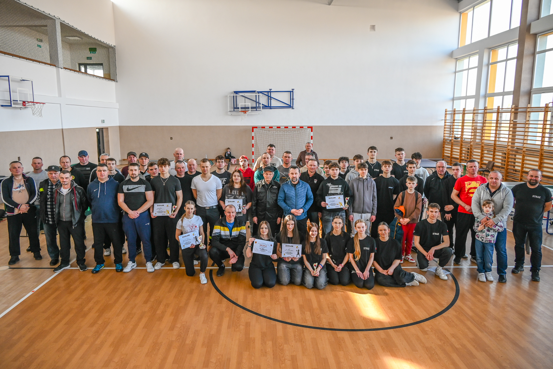 Duża grupa ludzi w hali sportowej na drewnianej podłodze, wśród nich osoby trzymające dyplomy i puchary, w tle bramka i kosze do koszykówki.