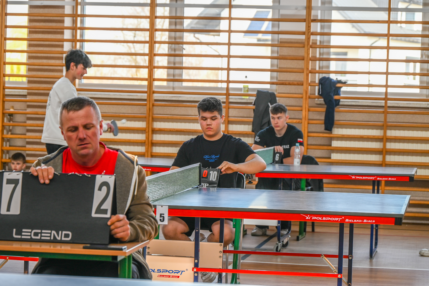 Mężczyzna w czerwonej koszulce i kurtce zmienia wynik 7:2 przy stole do tenisa stołowego, za nim dwóch młodych chłopców w czarnych koszulkach przy innych stołach.