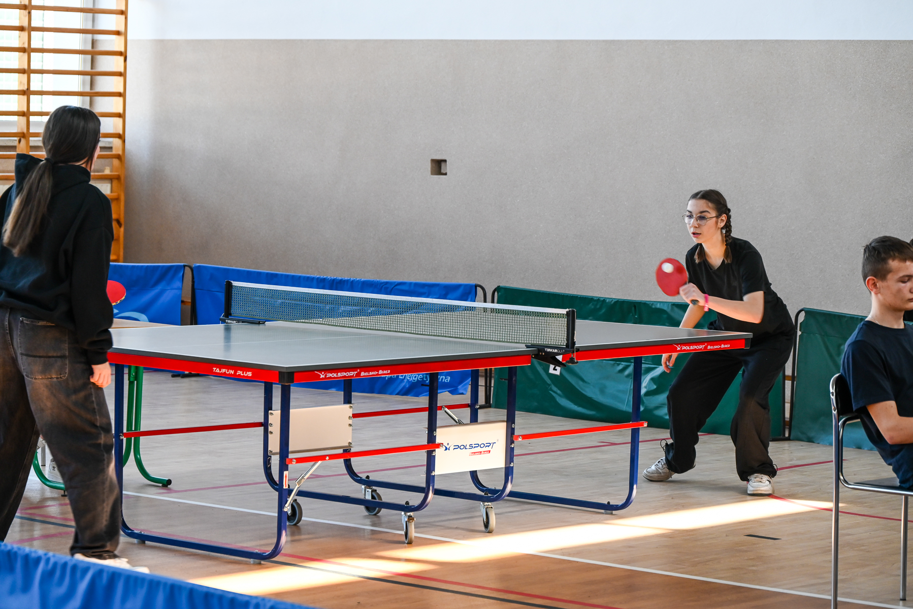 Dwie osoby grające w tenisa stołowego przy stole z siatką, jedna osoba siedzi na krześle po prawej stronie. W tle ściana i drewniane drabinki.