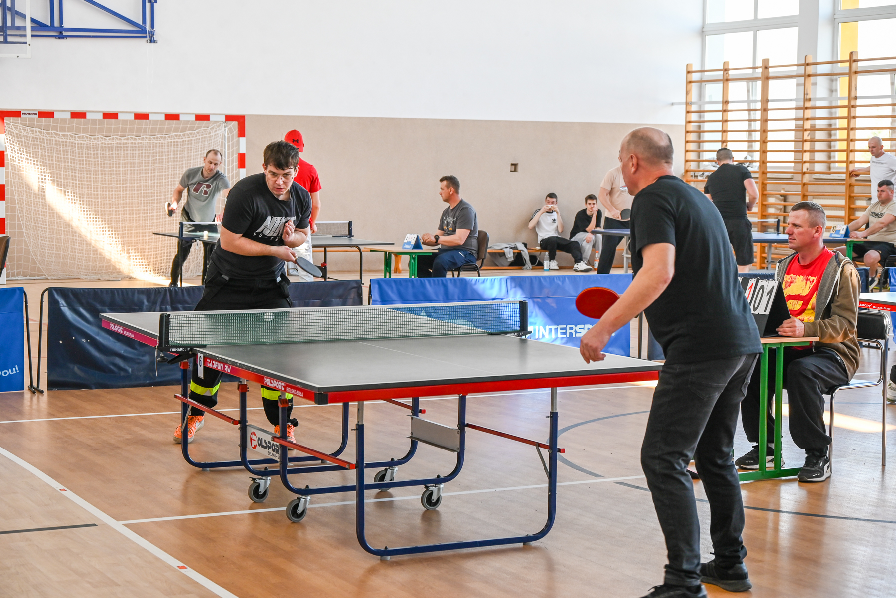 Dwóch mężczyzn gra w tenisa stołowego przy stole, w tle sędzia i inni obserwujący, jasna sala sportowa z drewnianą podłogą i oknami.