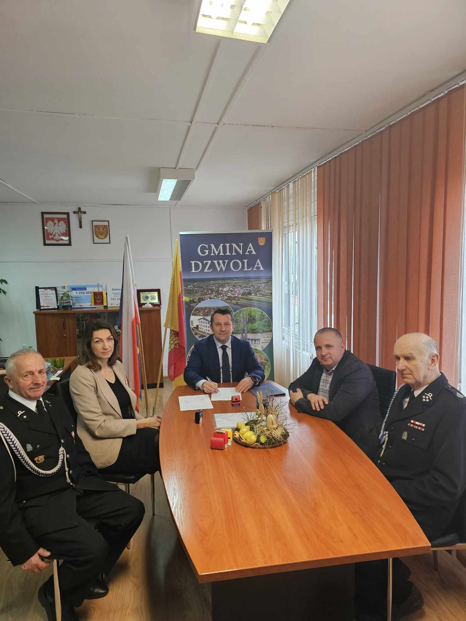 Pięć osób siedzi przy drewnianym stole w biurze z flagami i banerem "Gmina Dzwola", za nimi okna z pionowymi żaluzjami, na ścianie godło Polski i krzyż.