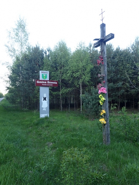 Drewniany krzyż z kwiatami w różnych kolorach na zielonej łące obok zielonego i czerwonego znaku z napisem Gmina Dzwola oraz lasu z drzew iglastych i liściastych w tle.