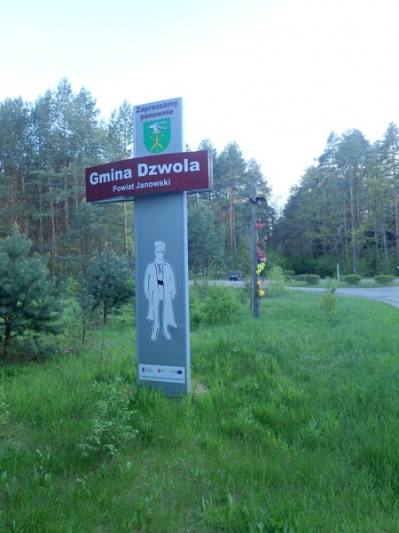 Tablica z napisem "Gmina Dzwola Powiat Janowski" na tle zielonej trawy i lasu z drzew iglastych, na słupie grafika postaci w stroju historycznym.