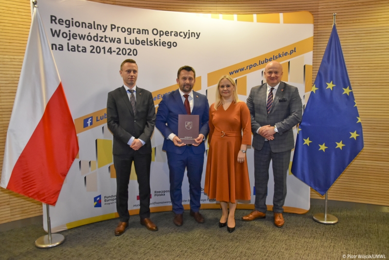 Cztery osoby w formalnych strojach stoją na tle baneru z napisem Regionalny Program Operacyjny Województwa Lubelskiego, po bokach flagi Polski i Unii Europejskiej.