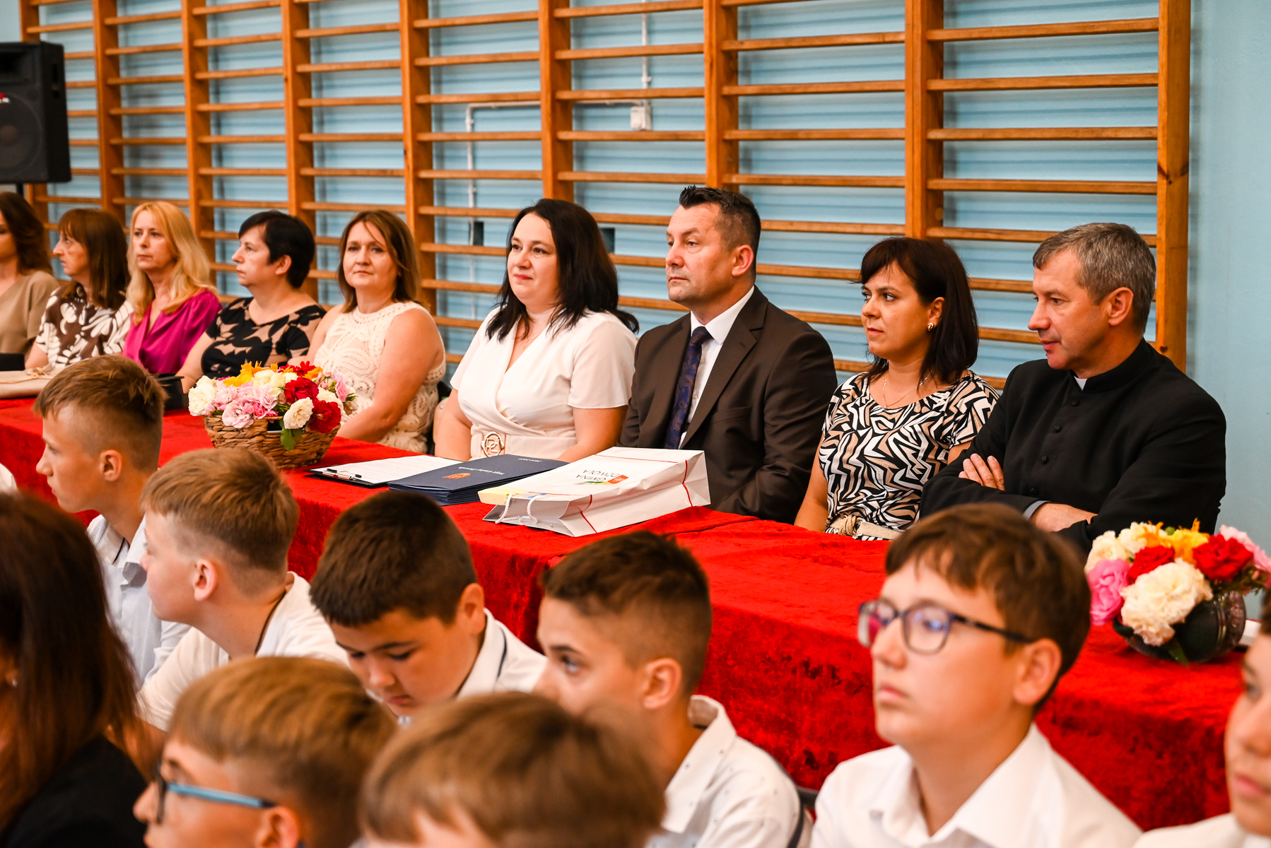 Grupa dorosłych siedzi za stołem z czerwonym obrusem, przed nimi dzieci w białych koszulach, w tle drewniane drabinki na niebieskiej ścianie, na stole kosze z kolorowymi kwiatami.