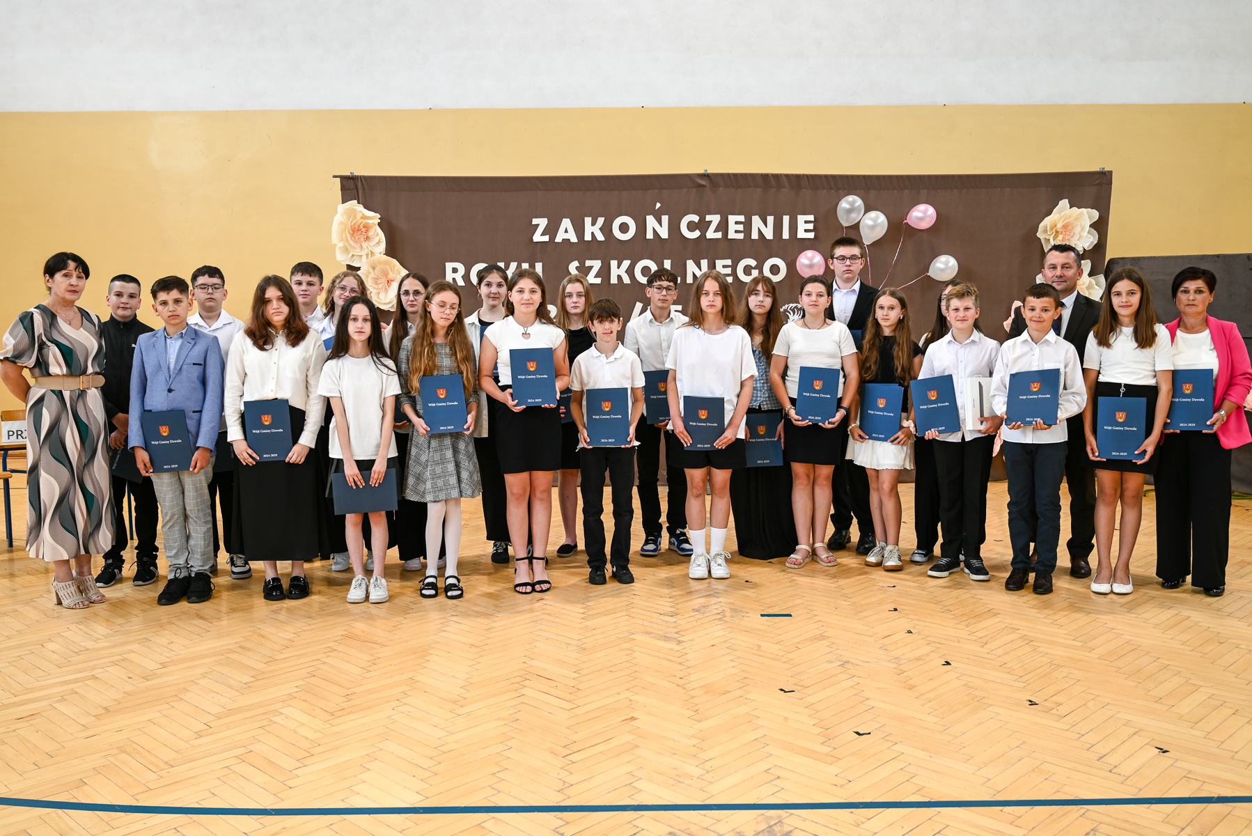 Grupa uczniów i nauczycieli na sali gimnastycznej, uczniowie trzymają granatowe teczki, za nimi brązowy baner z biały napisem i dekoracje kwiatowe.