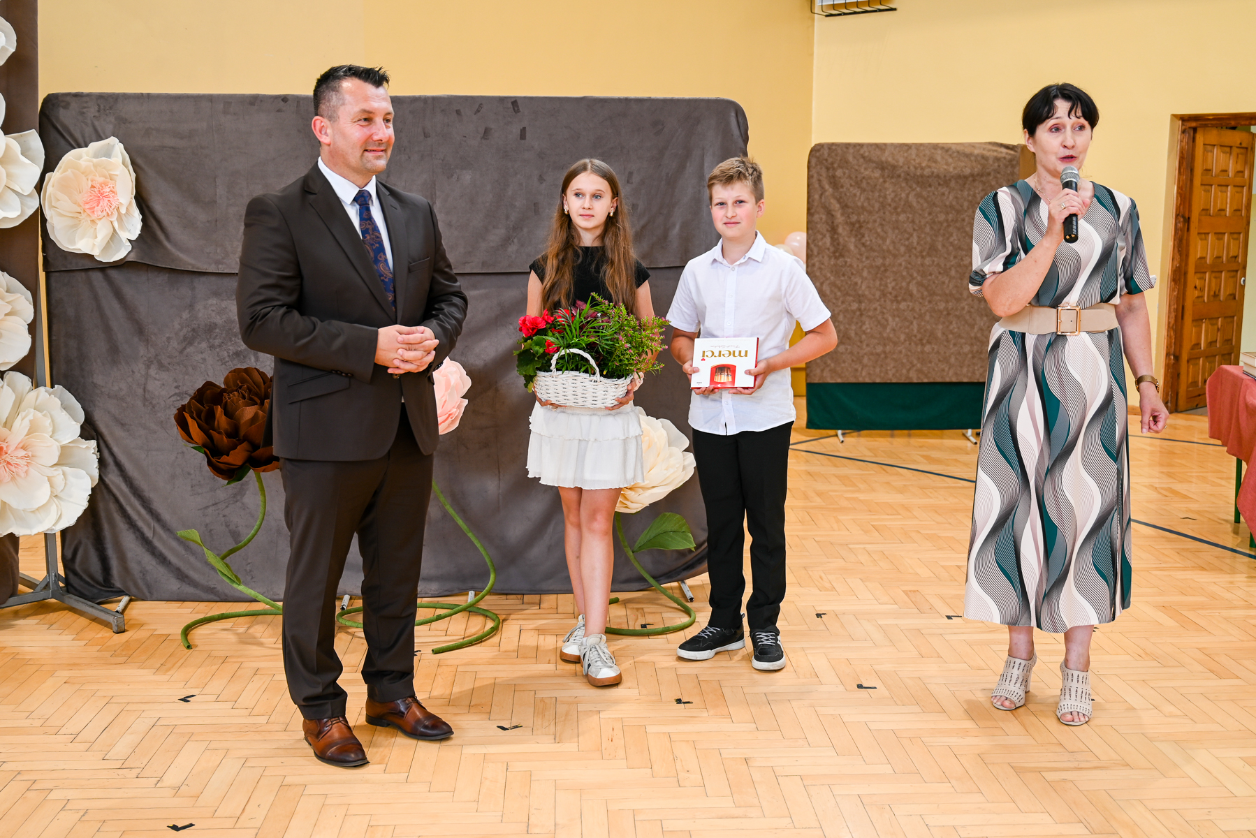 Mężczyzna w ciemnym garniturze, dziewczynka w białej spódnicy trzymająca kosz z kwiatami, chłopiec w białej koszuli z czekoladą, kobieta w sukience z mikrofonem na jasnej podłodze.