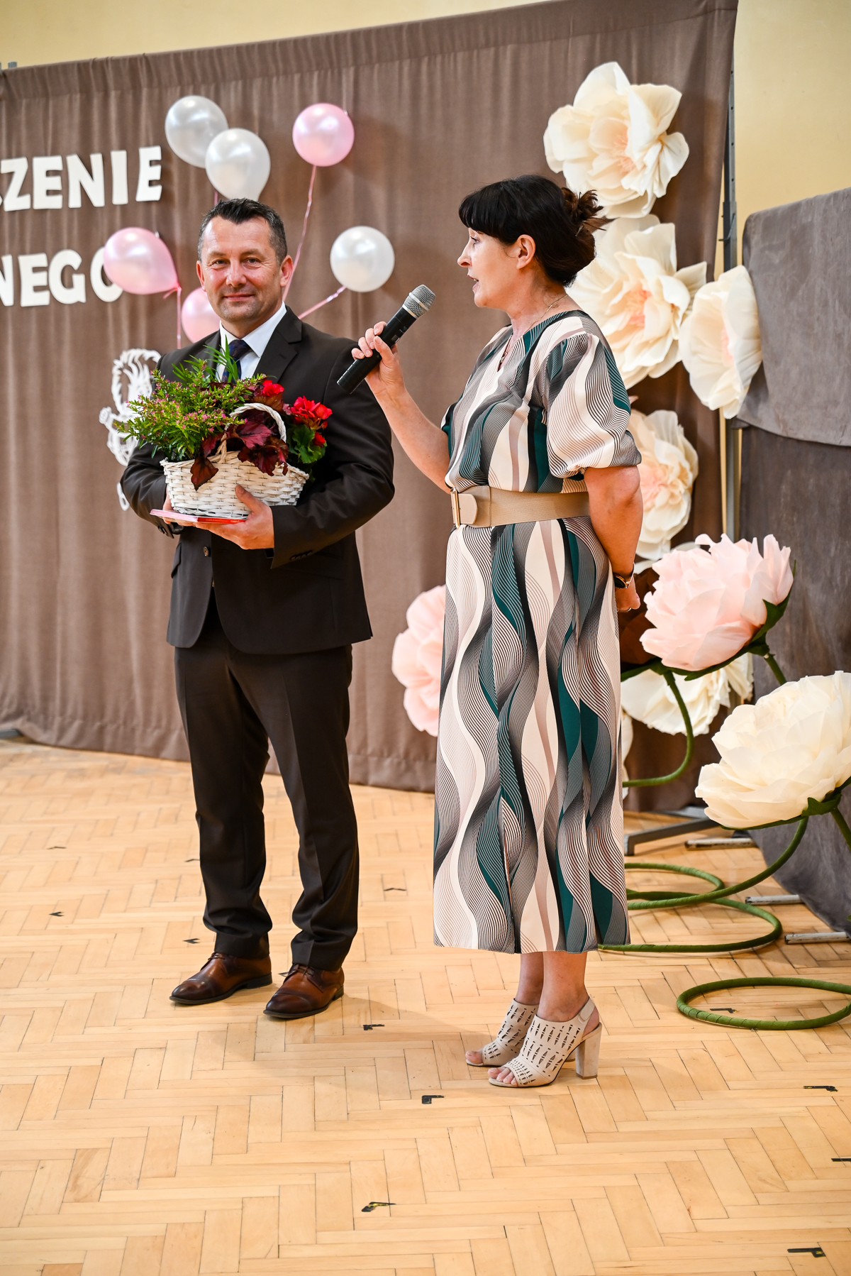 Mężczyzna w ciemnym garniturze trzyma kosz z kwiatami, kobieta w sukience w paski trzyma mikrofon, w tle balony i duże kremowe kwiaty na brązowej zasłonie.