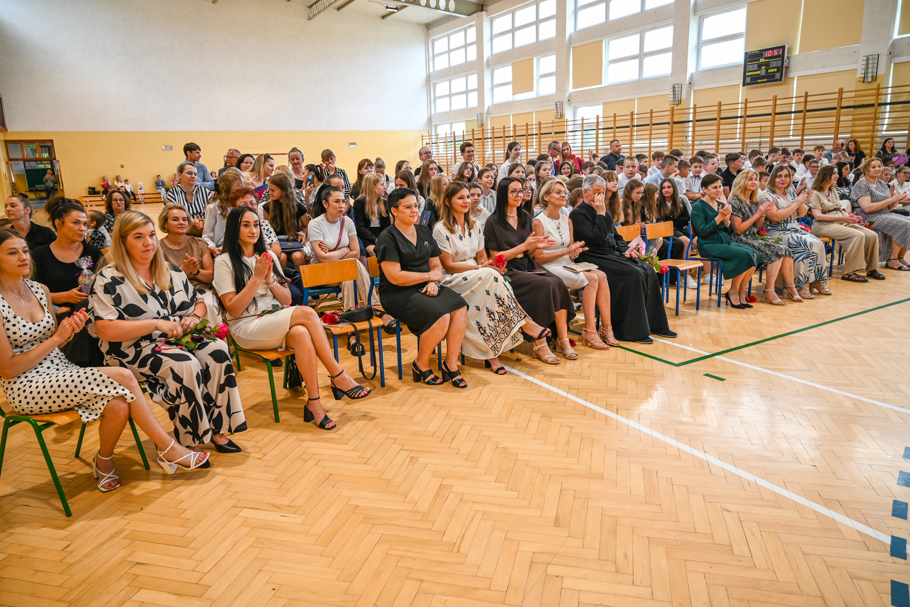 Wnętrze sali gimnastycznej z dużą grupą siedzących kobiet i młodzieży, drewniana podłoga, jasne ściany, duże okna, niektóre osoby trzymają czerwone róże.