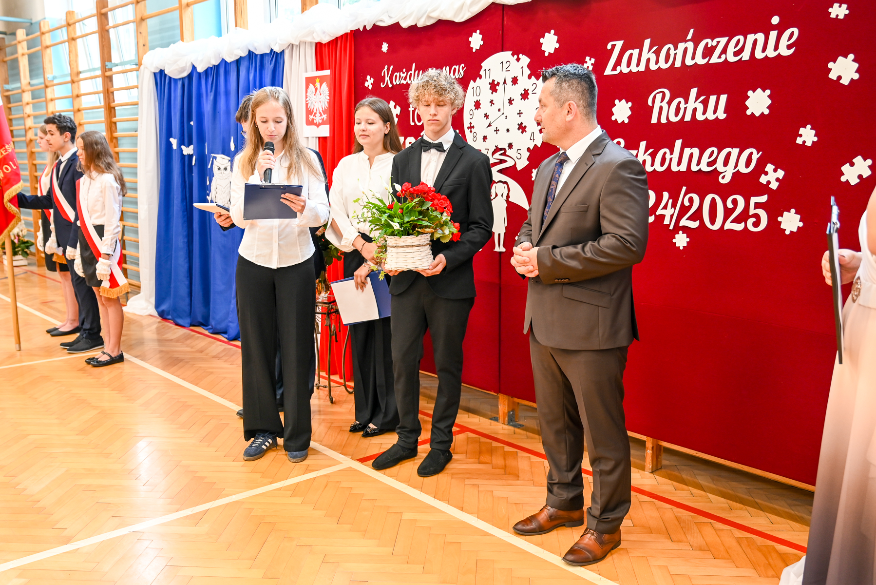 Grupa osób na sali gimnastycznej, dziewczyna z mikrofonem i kartką, chłopak w garniturze trzymający kosz z czerwonymi kwiatami, tło z czerwono-biało-niebieską dekoracją i napisem.
