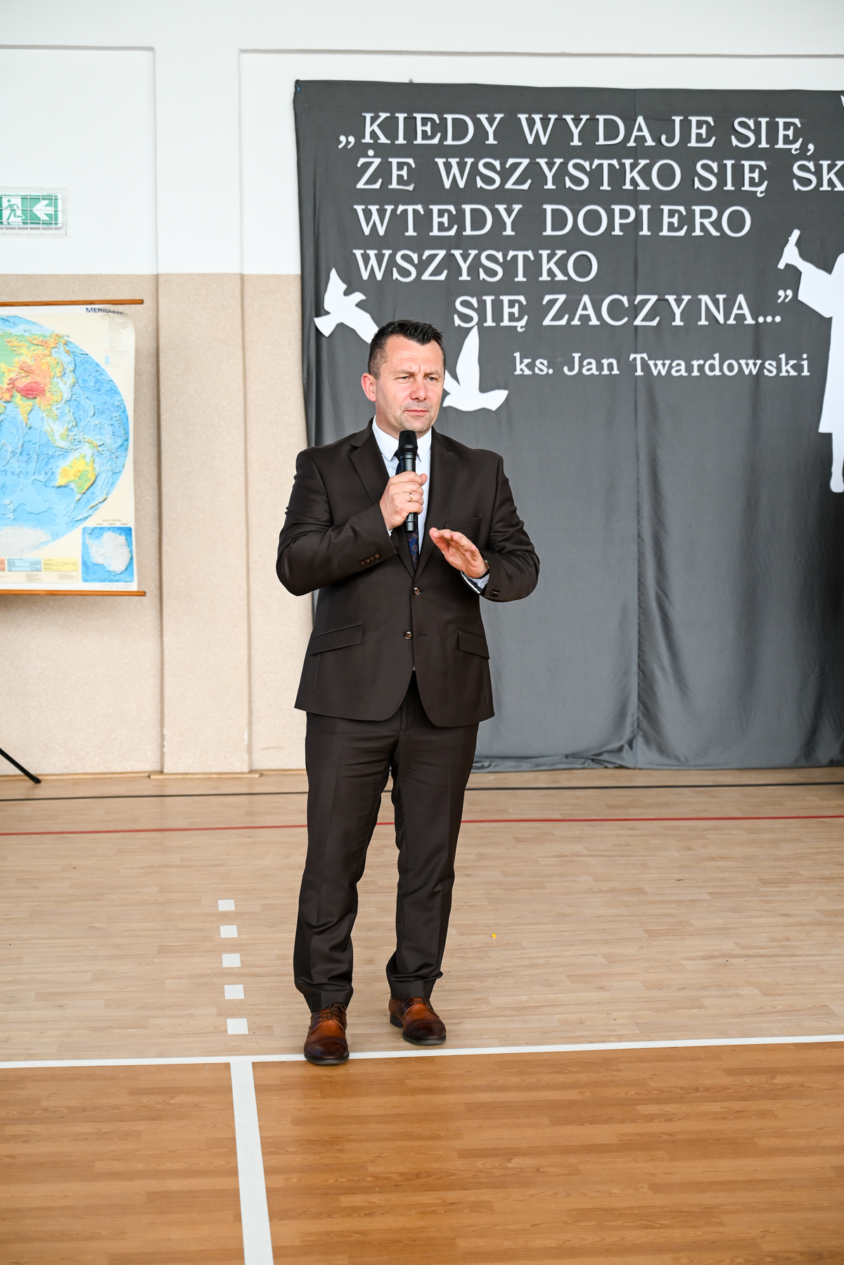 Mężczyzna w ciemnym garniturze i brązowych butach trzyma mikrofon, stoi na jasnej drewnianej podłodze, za nim szara kurtyna z białym napisem i sylwetkami ptaków.