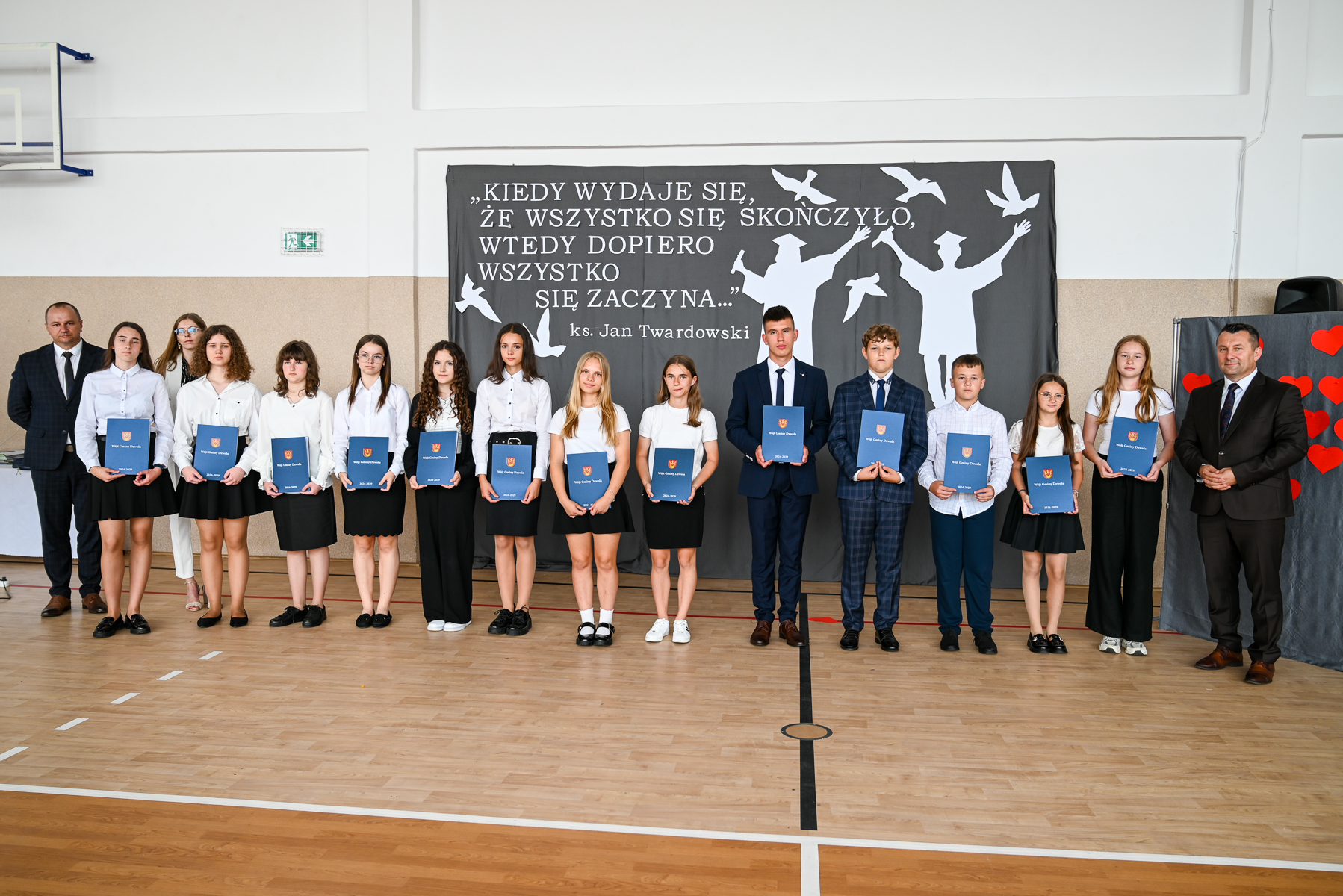 Grupa nastolatków i dwóch dorosłych stojących na tle szarego baneru z białym napisem i sylwetkami absolwentów oraz ptaków, trzymających niebieskie teczki, w jasnym pomieszczeniu z drewnianą podłogą.