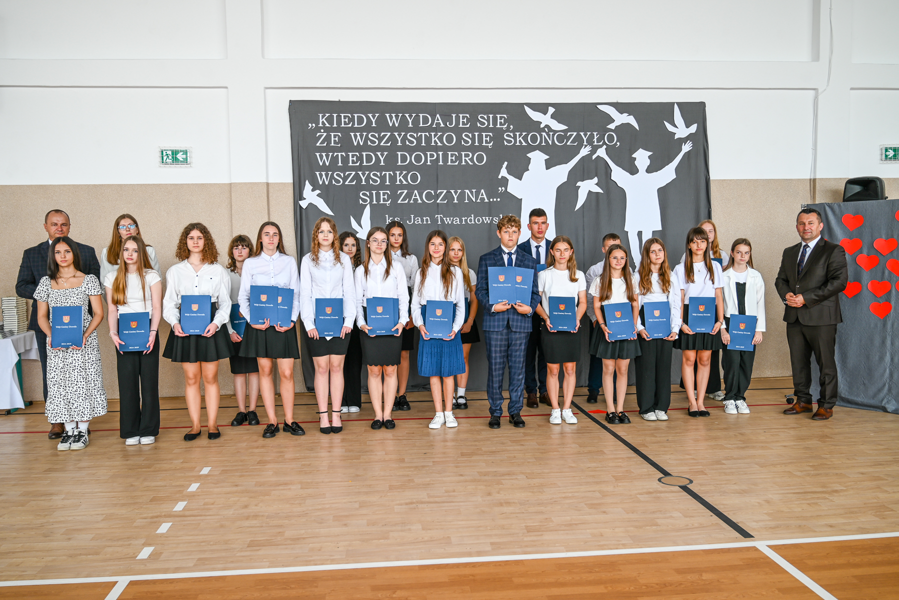 Grupa młodzieży i dwóch mężczyzn na tle czarnego baneru z białym napisem i sylwetkami ptaków oraz postaci absolwentów, trzymają niebieskie teczki, w sali z jasną podłogą.