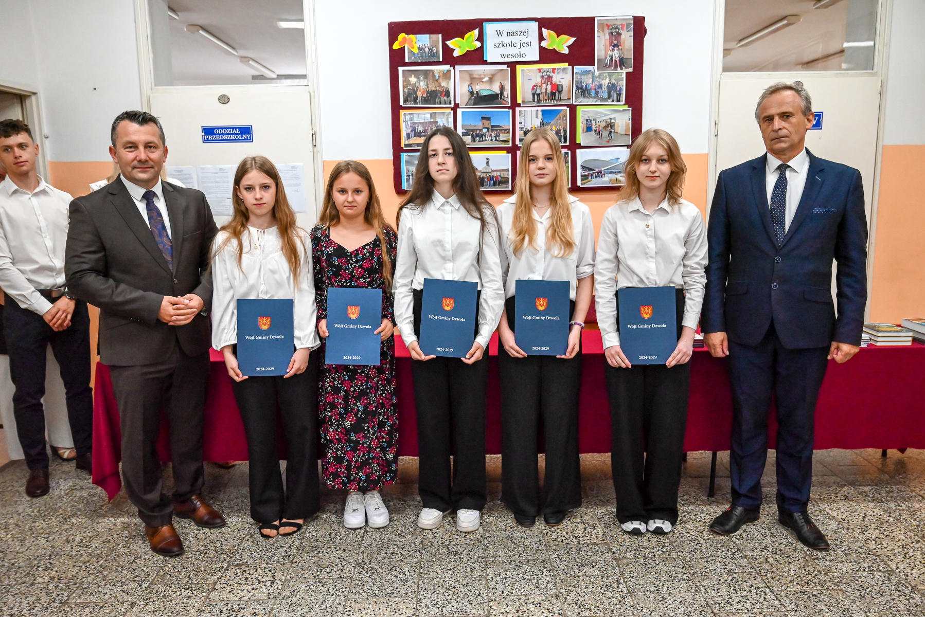 Grupa osób stojących na korytarzu szkolnym, w tym pięć dziewcząt trzymających granatowe teczki oraz dwóch mężczyzn w garniturach po bokach. Za nimi tablica ze zdjęciami.