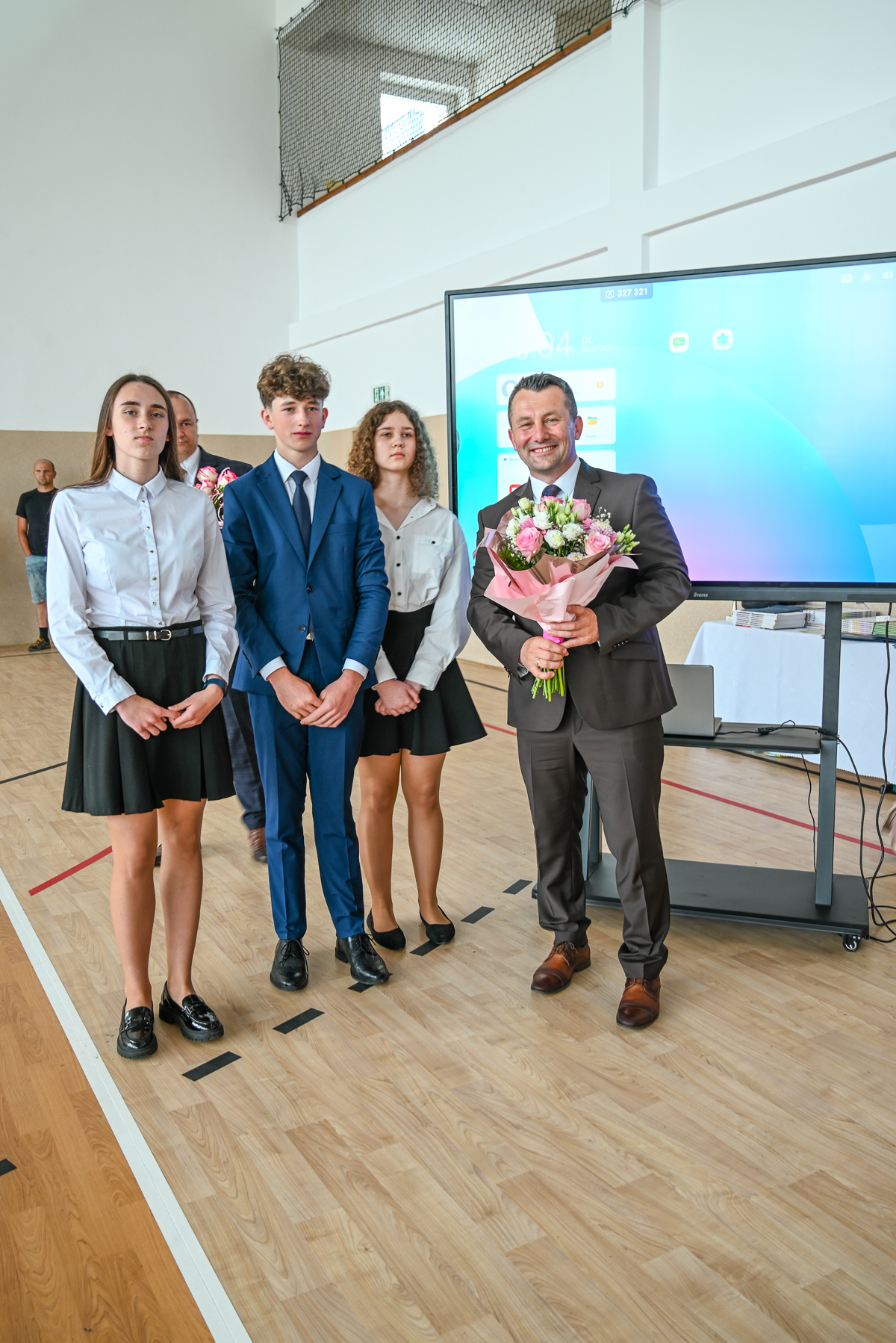 Trójka młodych ludzi w szkolnych strojach, mężczyzna w garniturze z bukietem kwiatów, za nimi ekran dotykowy i drewniana podłoga sali gimnastycznej.